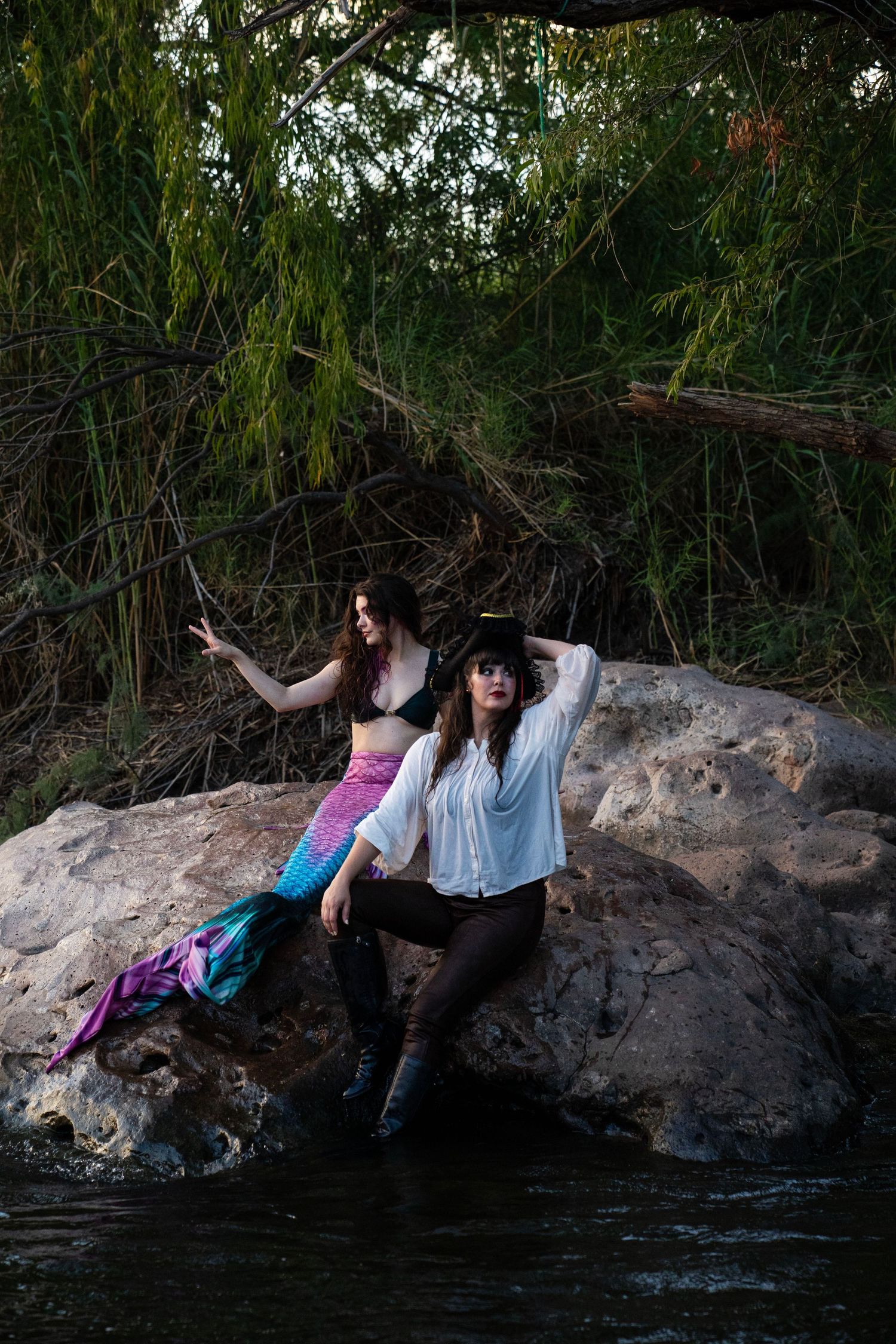 Two women in mermaid and pirate costumes pose on rocks by a river at dusk. Mermaid photoshoot, fantasy photoshoot. Arizona photographer.