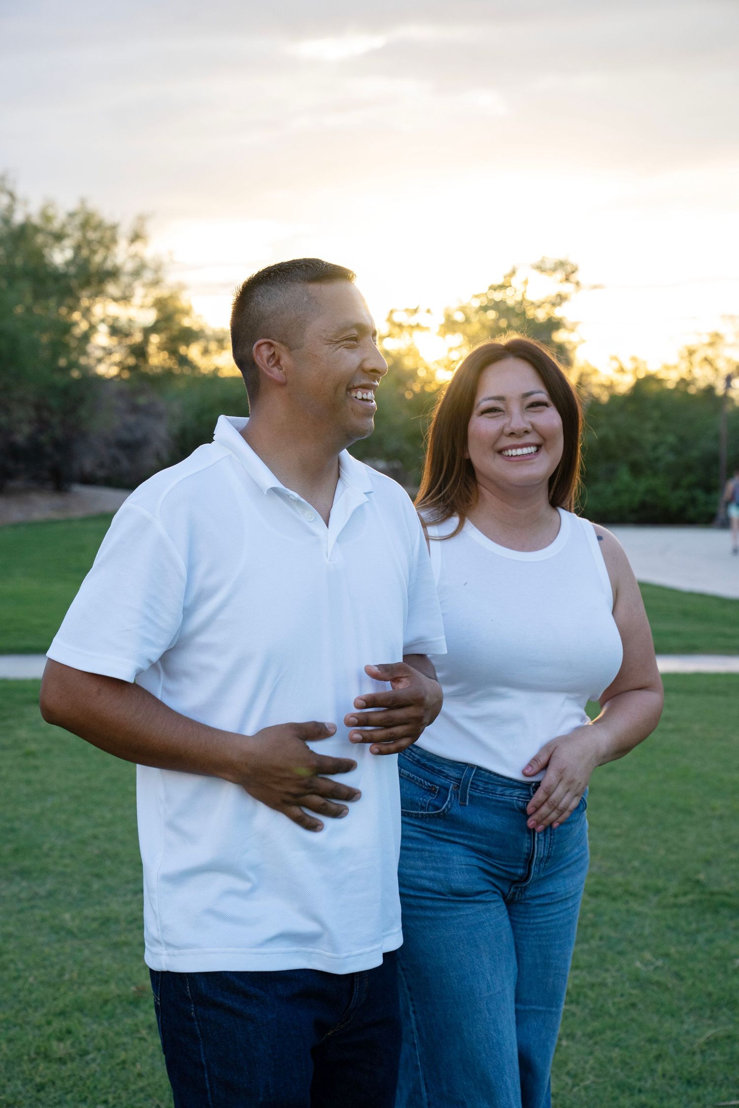 A smiling couple in matching white tops and jeans embrace outdoors at sunset with green trees in the background.