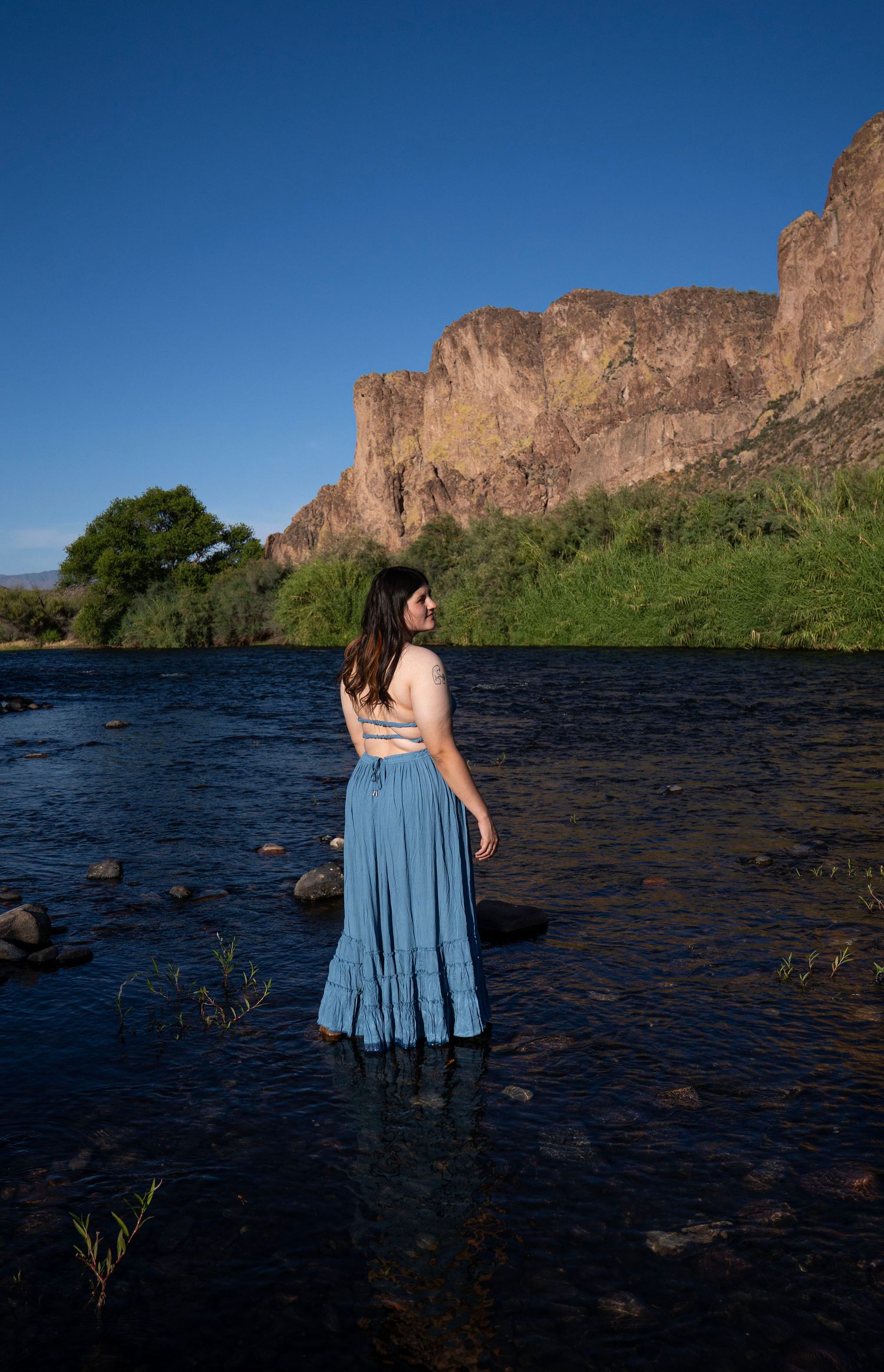 Woman in blue maxi skirt standing in a river with dramatic rocky cliffs and green trees under a clear blue sky.