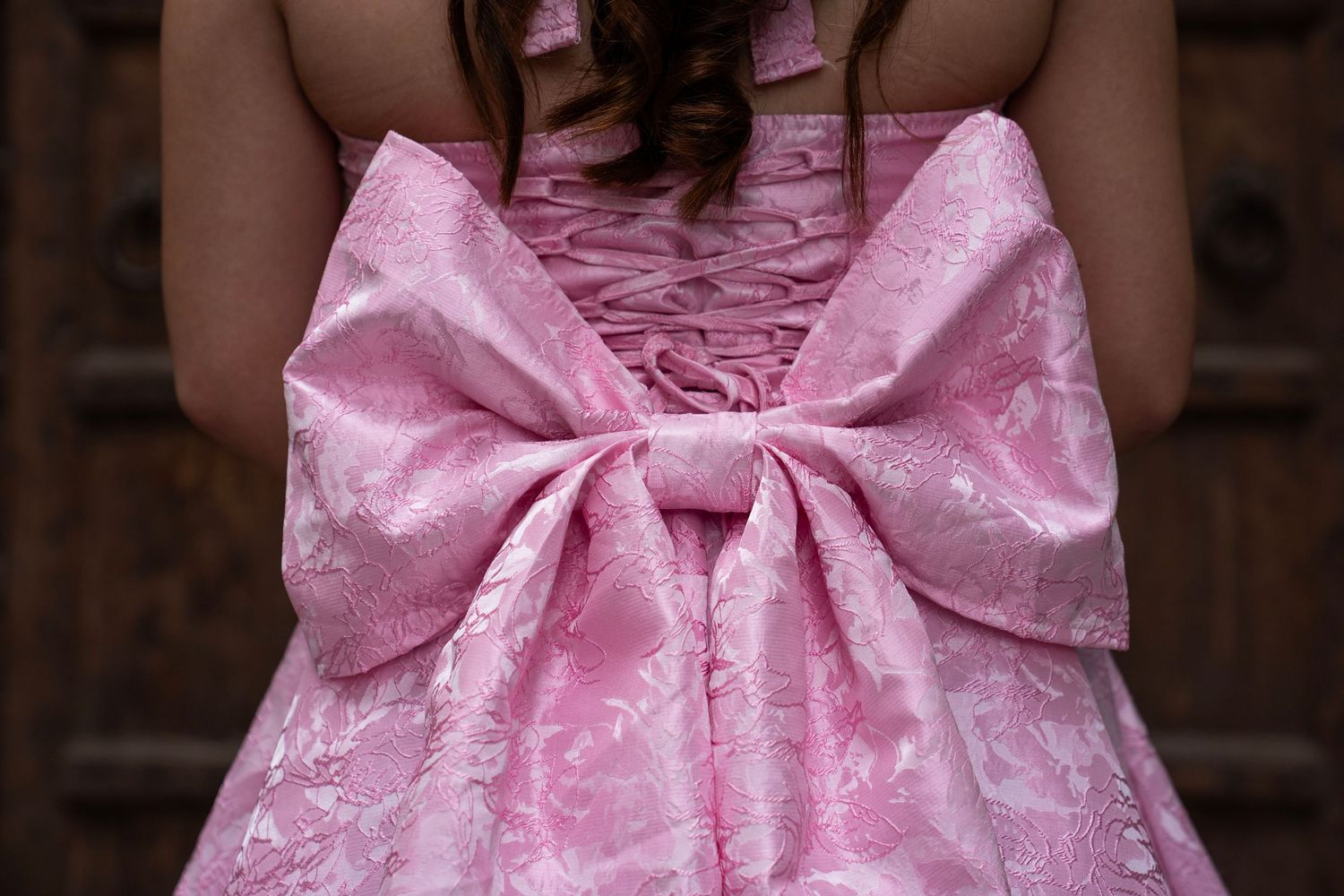 Young woman in elegant pink brocade dress with large bow detail on the back, photographed from behind.