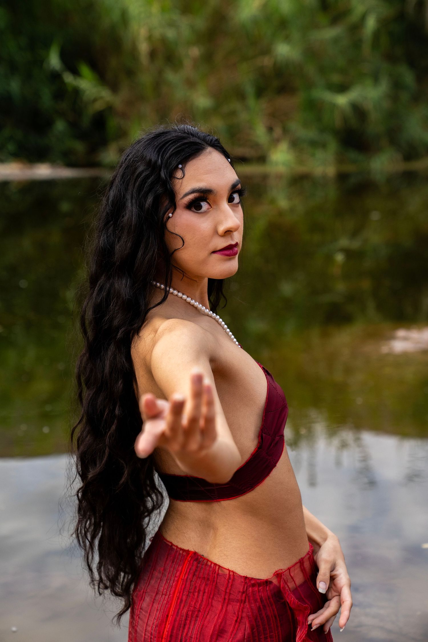 A woman in a burgundy bralette and red skirt reaches toward the camera while standing in a reflective outdoor river.