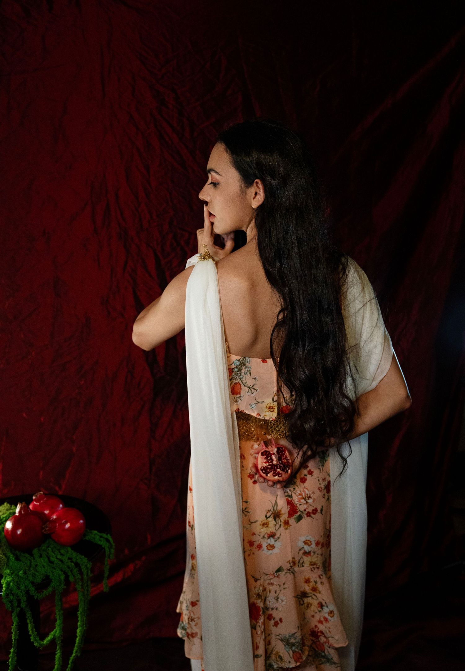 Woman with long dark hair in a floral dress and white shawl holds a finger to her lips before a red velvet backdrop beside red pomegranates.