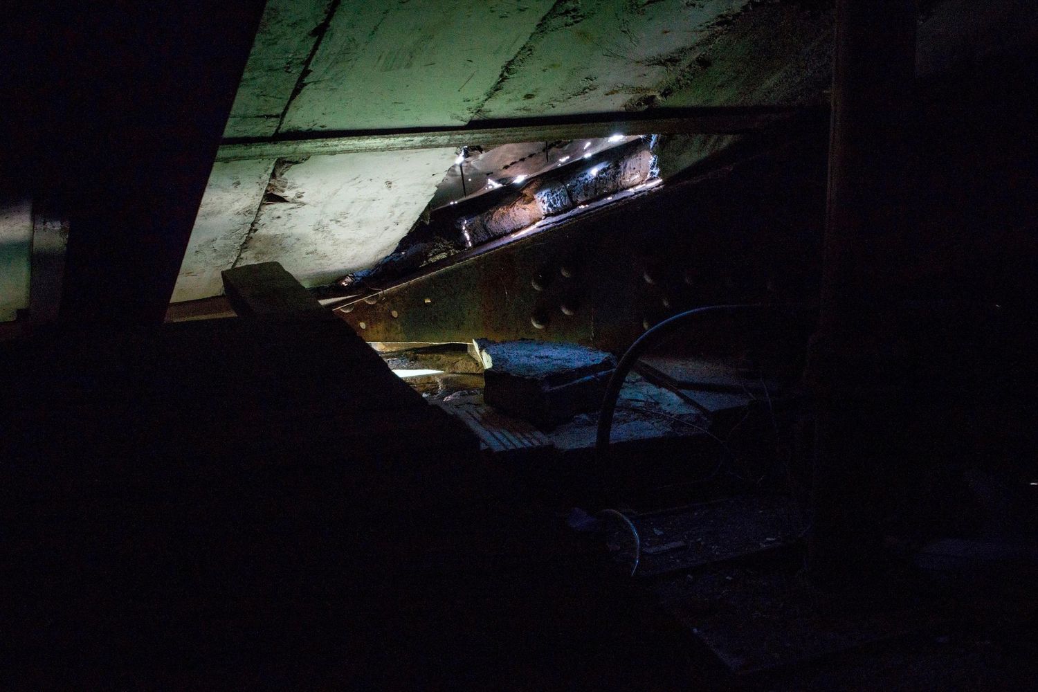 Dimly lit attic or crawl space with sloped wooden ceiling panels, exposed beams, and faint light glowing across cluttered shadowed surfaces.