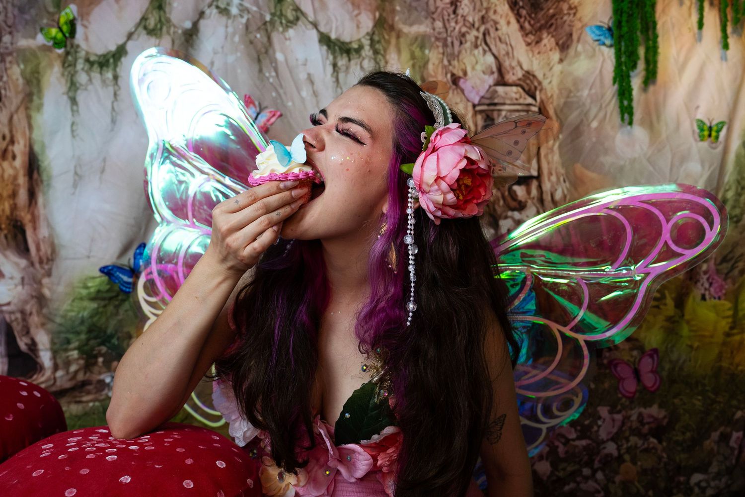Woman dressed as a fairy with butterfly wings and floral hair accessories eating a colorful cupcake. Fantasy Photoshoot