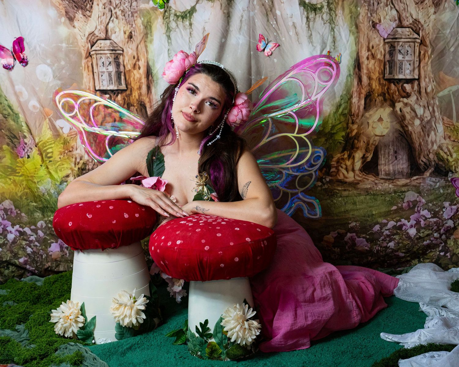 Model in pink fairy costume with multicolored wings poses with giant red mushrooms in magical forest setting. Fantasy Photoshoot