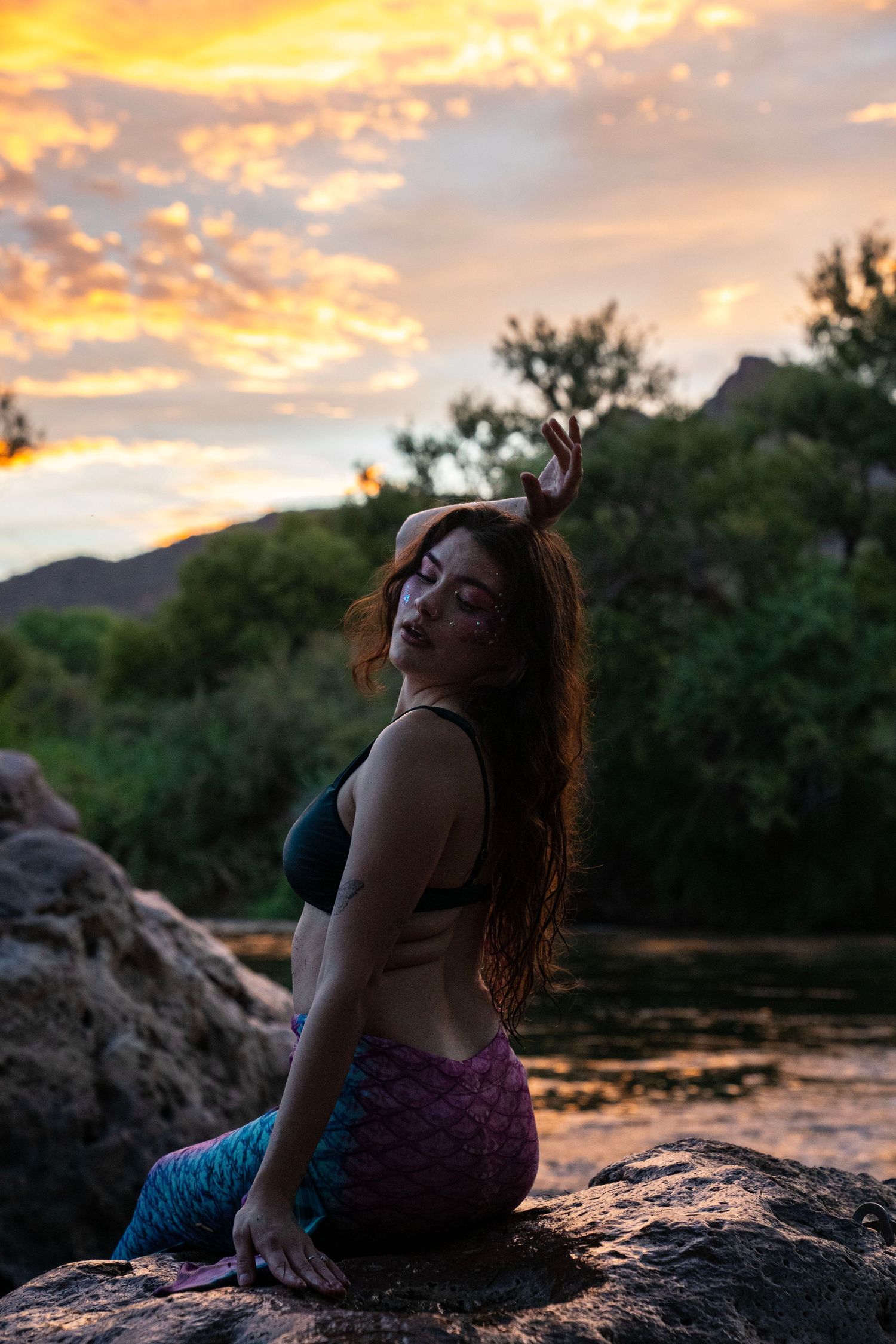 A woman wearing a colorful mermaid tail sits on rocks posing against a glowing orange sunset sky. Mermaid photoshoot, fantasy photoshoot.