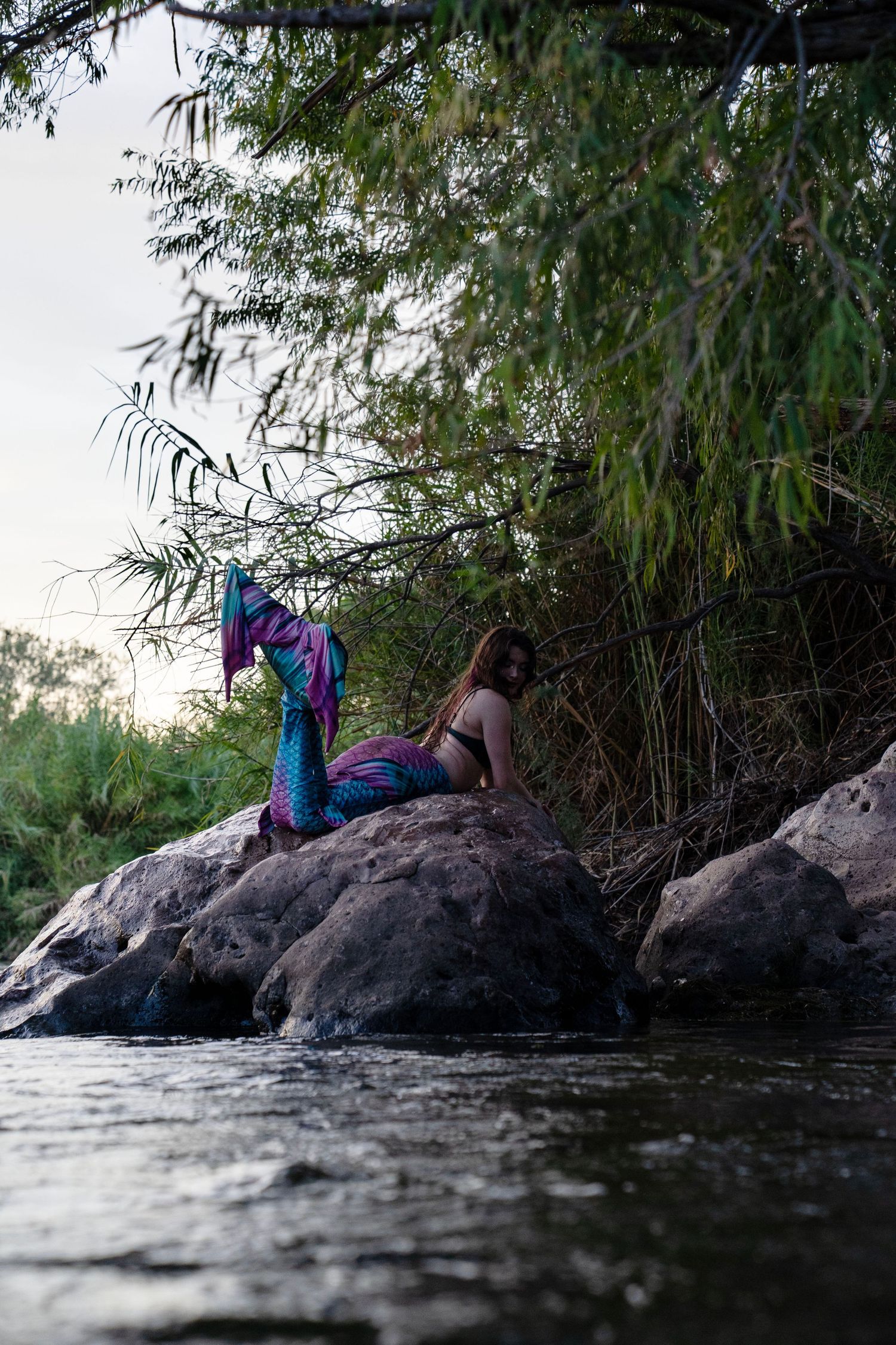 A woman in a colorful mermaid tail costume rests on rocks beside a flowing river surrounded by green foliage. Fantasy Photoshoot