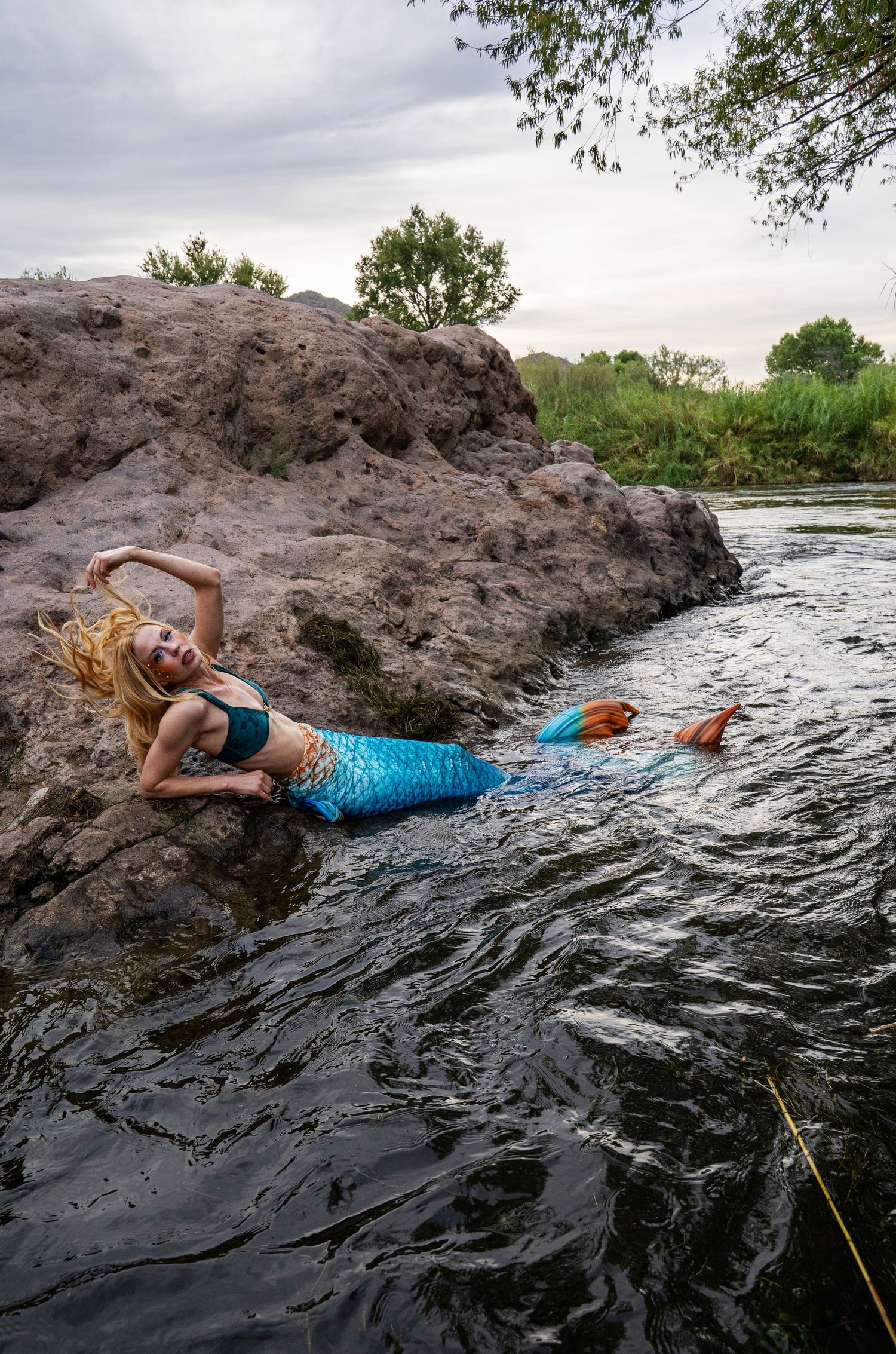A blonde woman in a teal mermaid costume with blue tail poses on rocks by a river at dusk. Fantasy Photoshoot