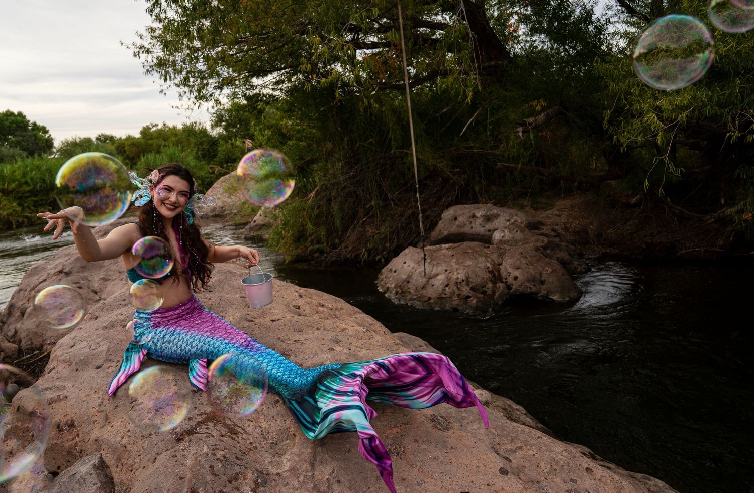 Wide shot of woman in colorful mermaid costume surrounded by large soap bubbles on rocky riverbank. Fantasy Photoshoot