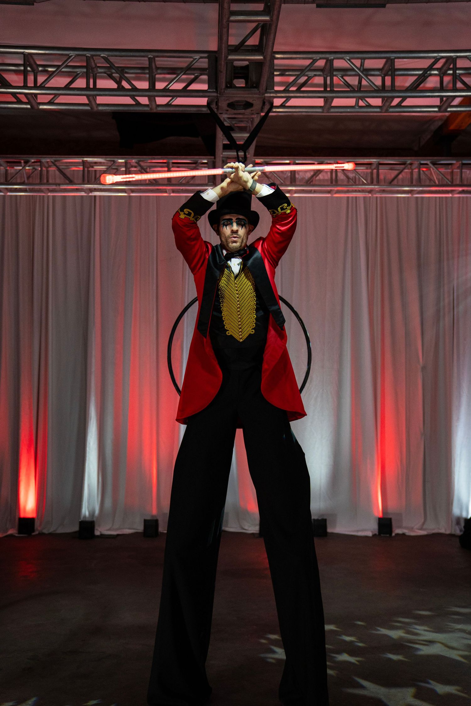 Circus ringmaster on stilts raises glowing batons overhead in a dramatic pose on a lit stage.
