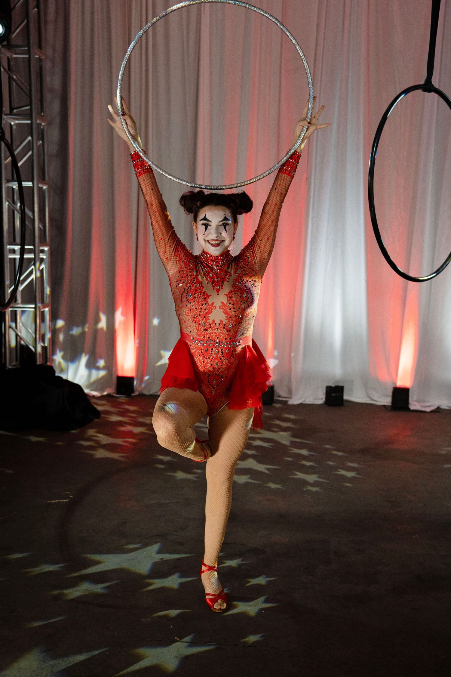 Circus performer in red sequin costume raises silver hoop overhead with clown makeup on stage.
