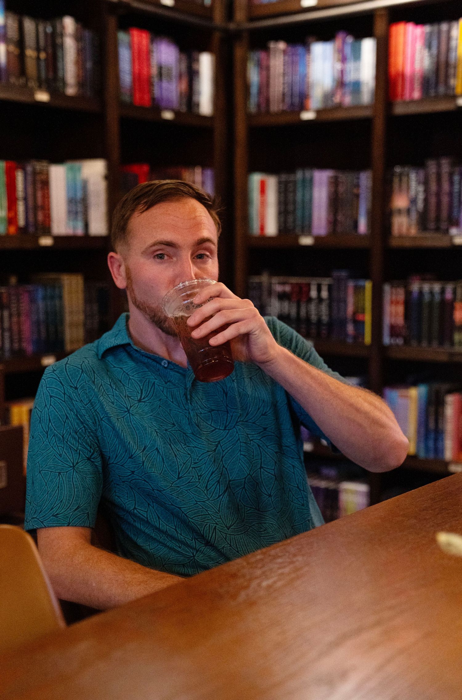 Man drinking beer at wooden table in front of bookshelf-lined library wall.