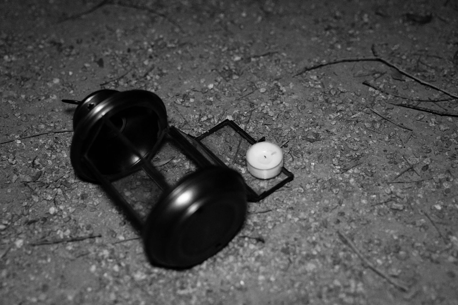 Black and white photo of a disassembled vintage lantern with a small lit candle resting on gravel and dry leaves on the ground.