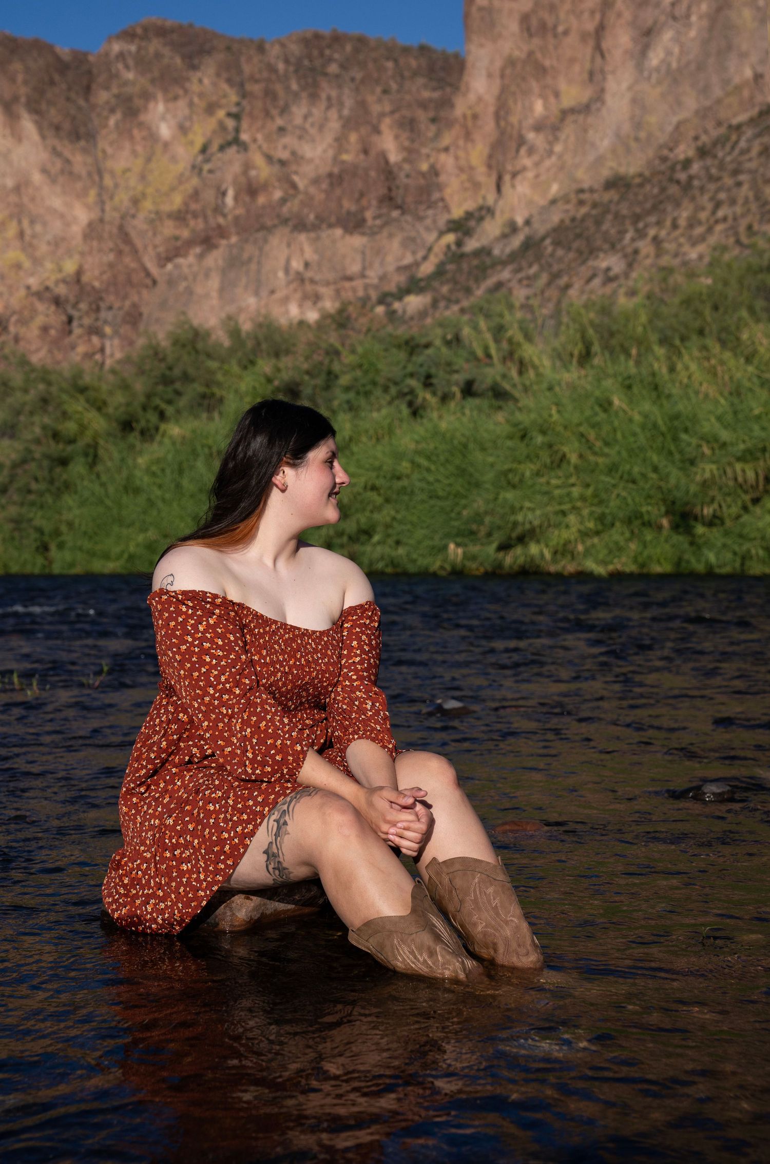 Woman in red floral off-shoulder dress sitting in a river with rocky mountains and green trees in background.