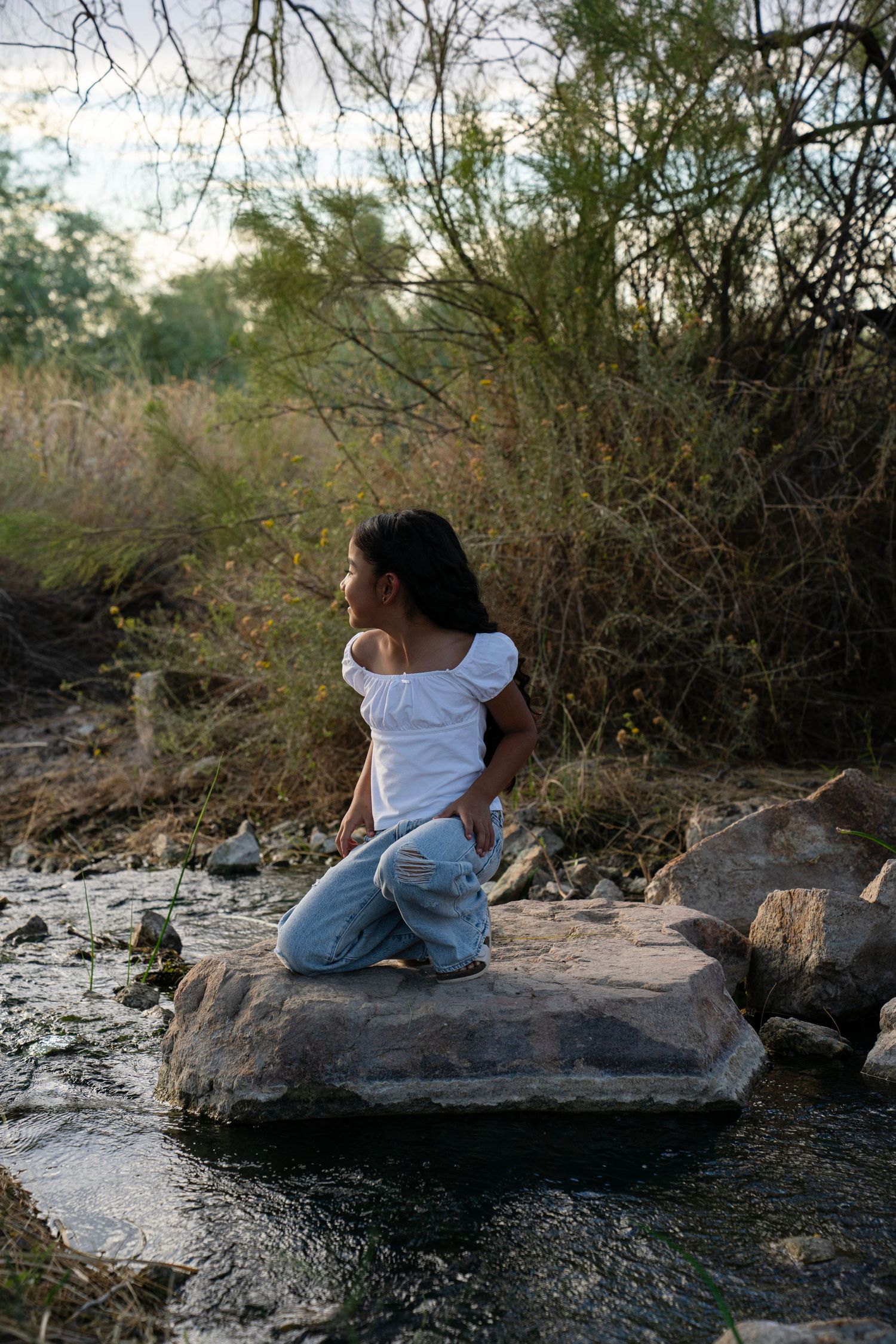 Young woman in white top and jeans sits peacefully on rocks beside a serene stream surrounded by lush greenery.