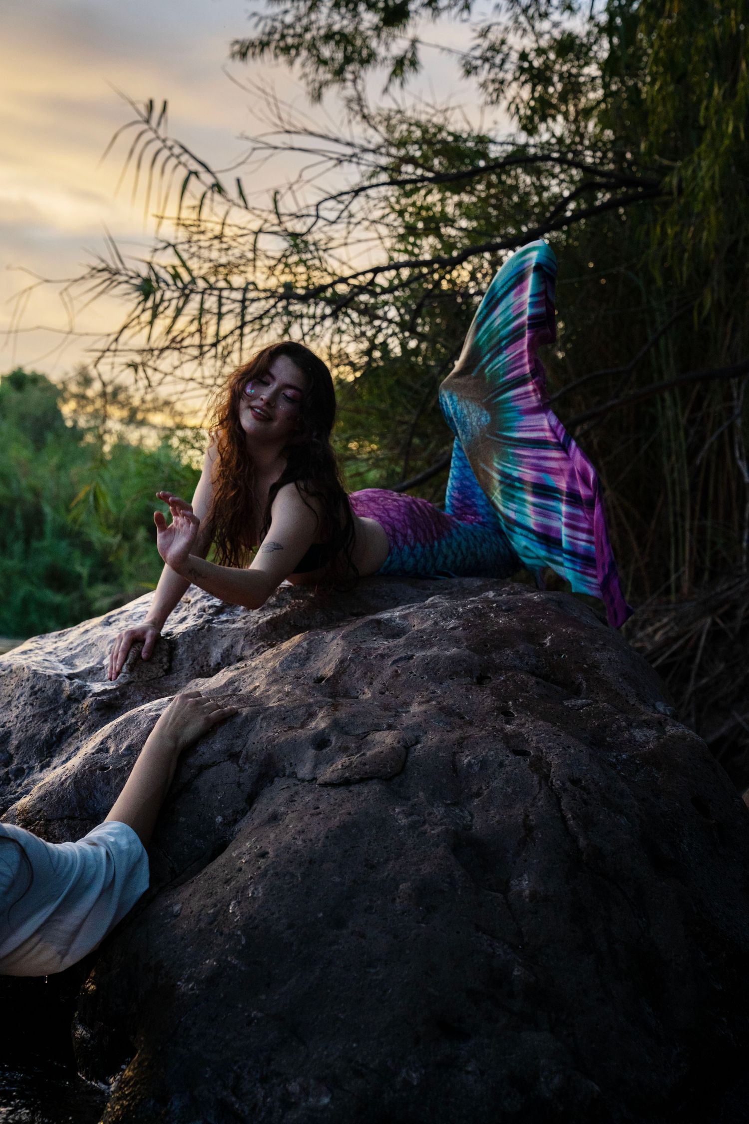 Woman in colorful mermaid tail costume reclines on rock near river at sunset. Mermaid photoshoot, fantasy photoshoot. Arizona photographer.