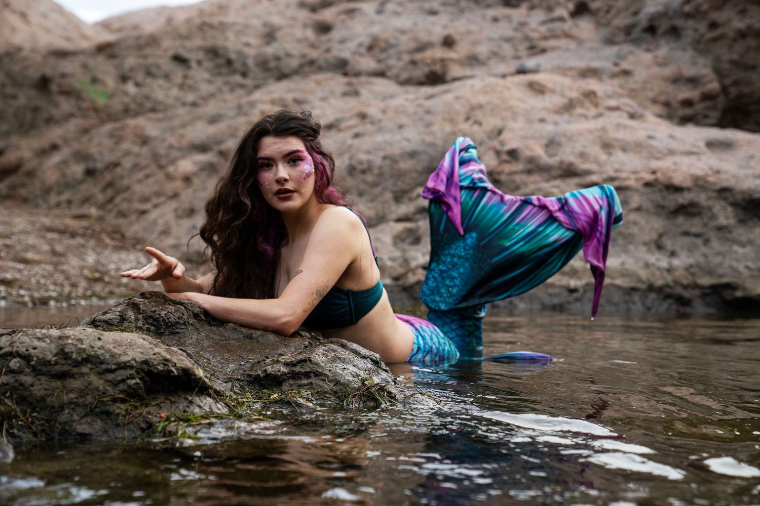 A woman in mermaid cosplay with pink hair and colorful tail poses on rocks in shallow water. Fantasy Photoshoot