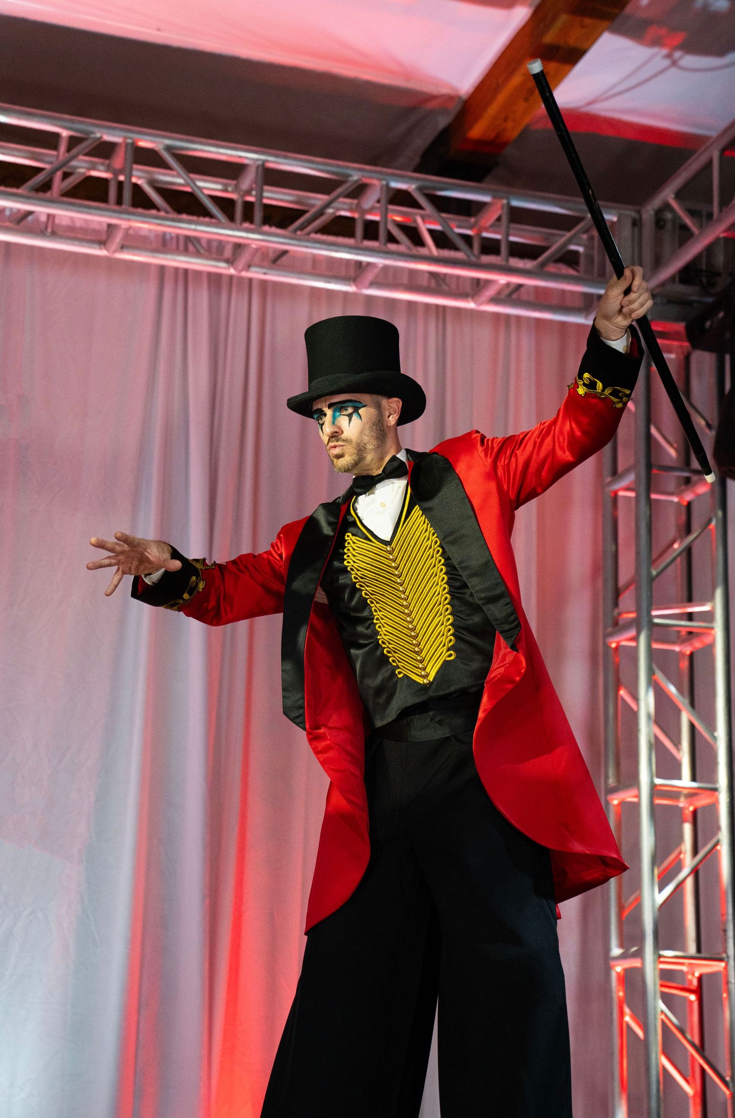 Male ringmaster in red tailcoat and top hat performs on stage with dramatic pose and baton.