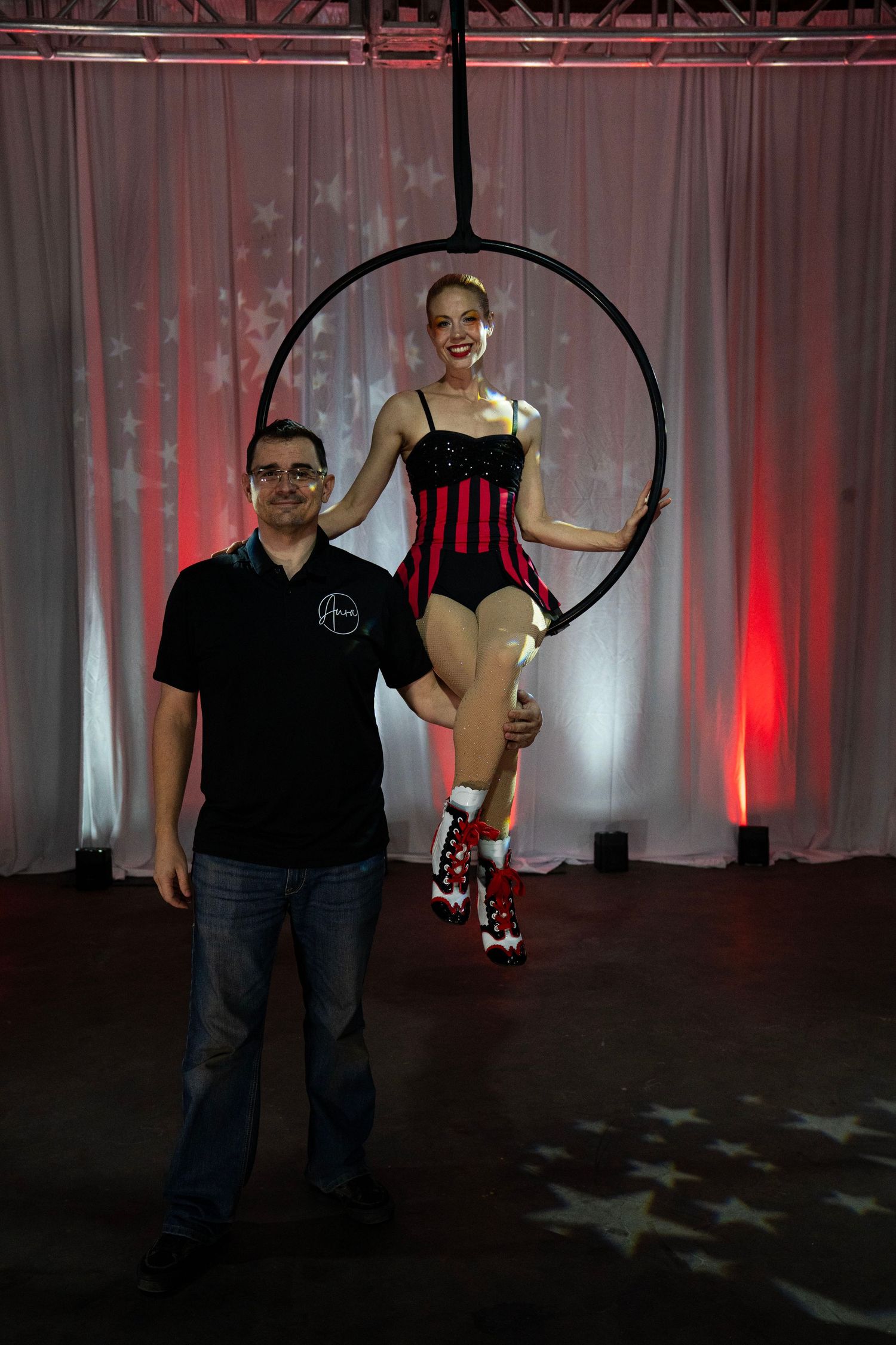 A smiling aerial hoop performer in red and black costume poses with a man in black shirt at a circus event.