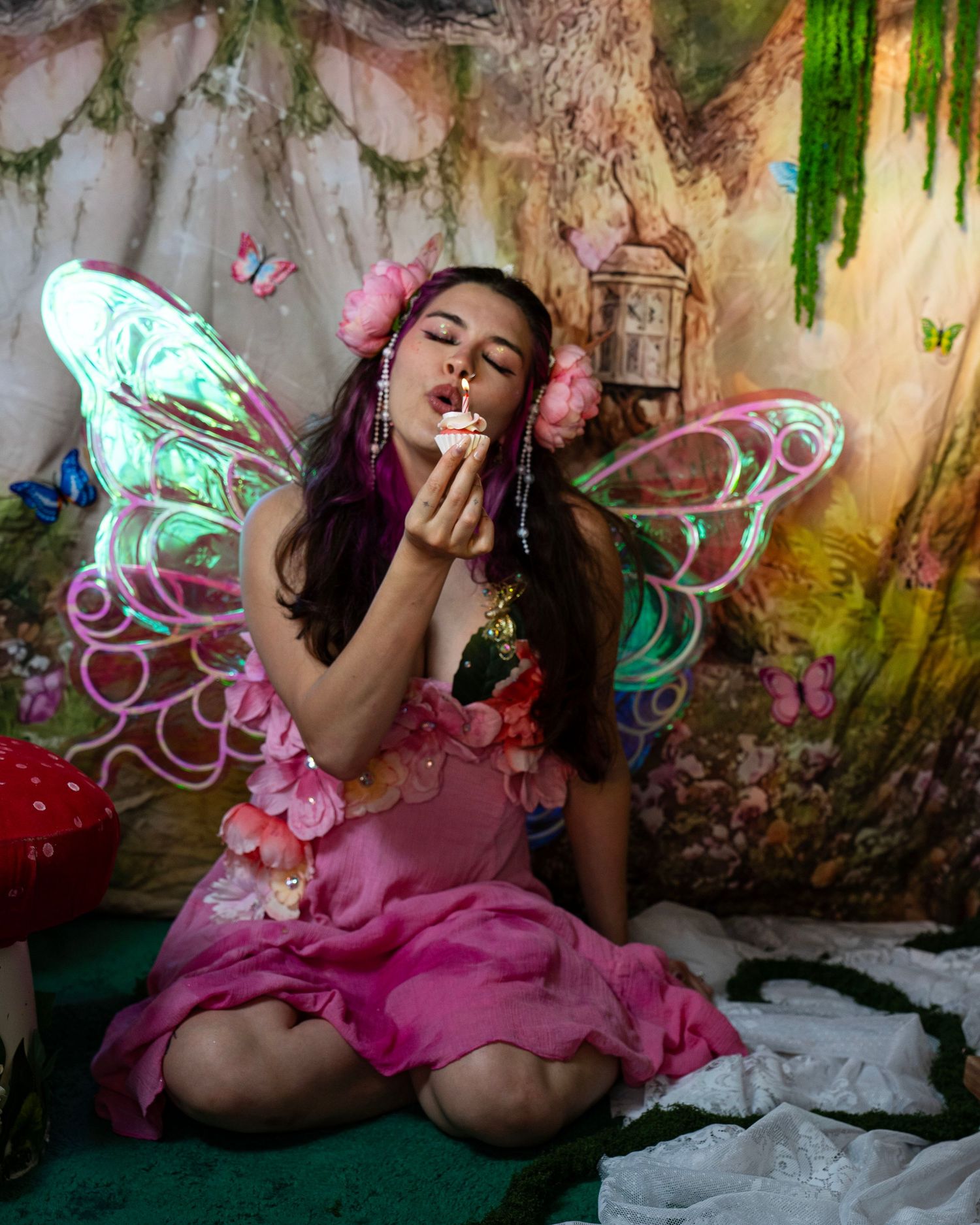 Fairy in pink floral dress with iridescent wings blows a kiss while holding a cupcake in an enchanted forest. Fantasy Photoshoot