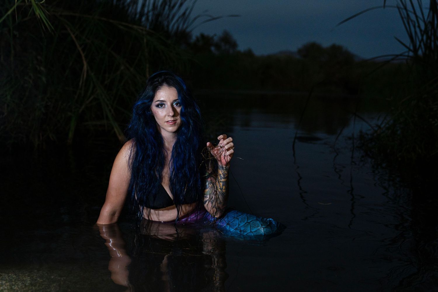 Woman with long blue hair in mermaid tail posing in dark lake at dusk.