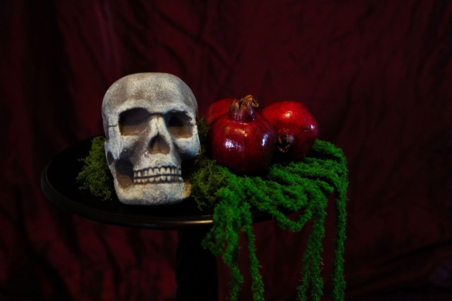 Human skull beside ripe red pomegranates on a dark stand with trailing green moss against a deep red velvet backdrop.