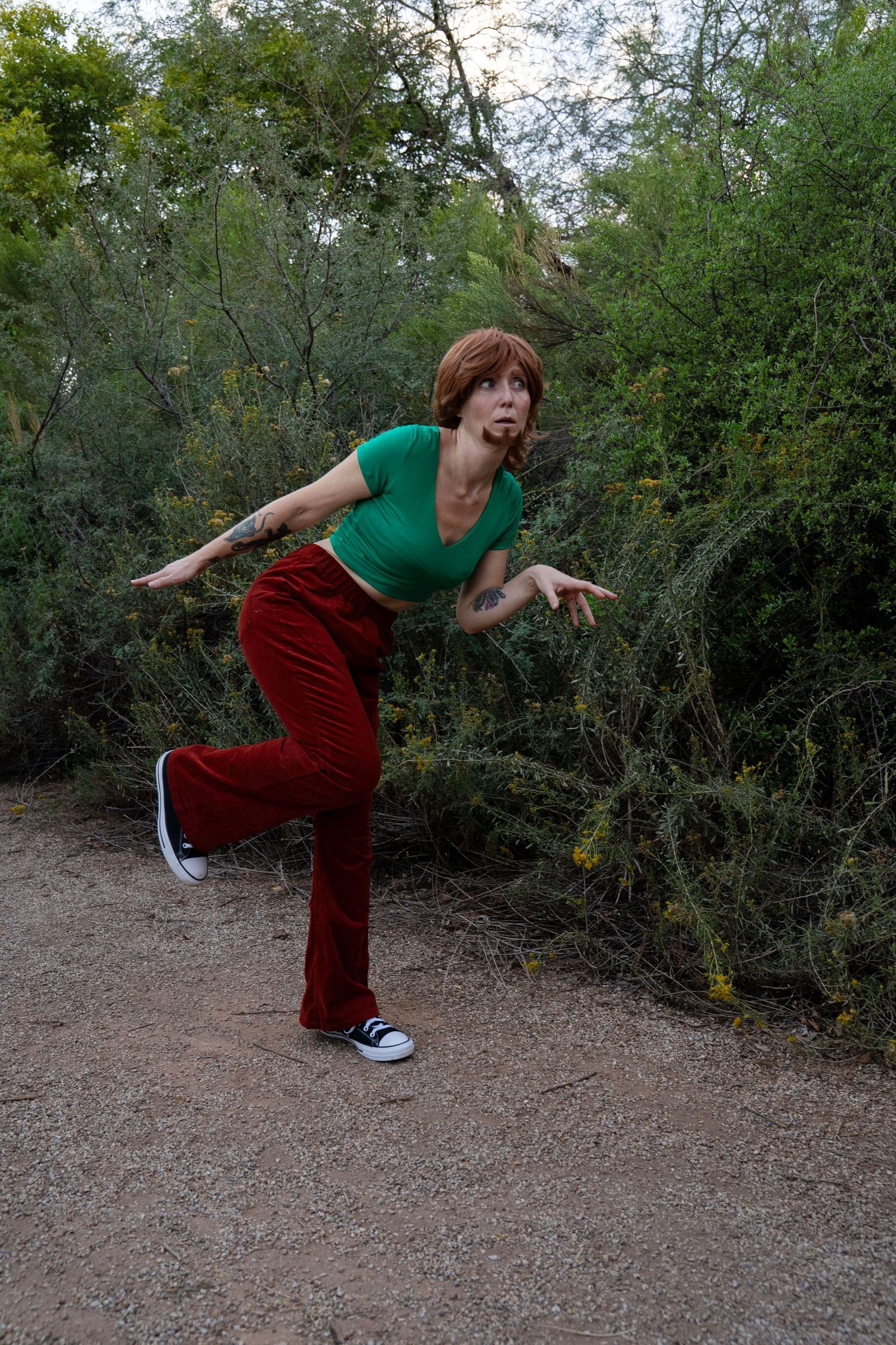 Person in a green crop top, red flared pants and black sneakers strikes a playful crouching pose on a dirt path beside green desert shrubs.