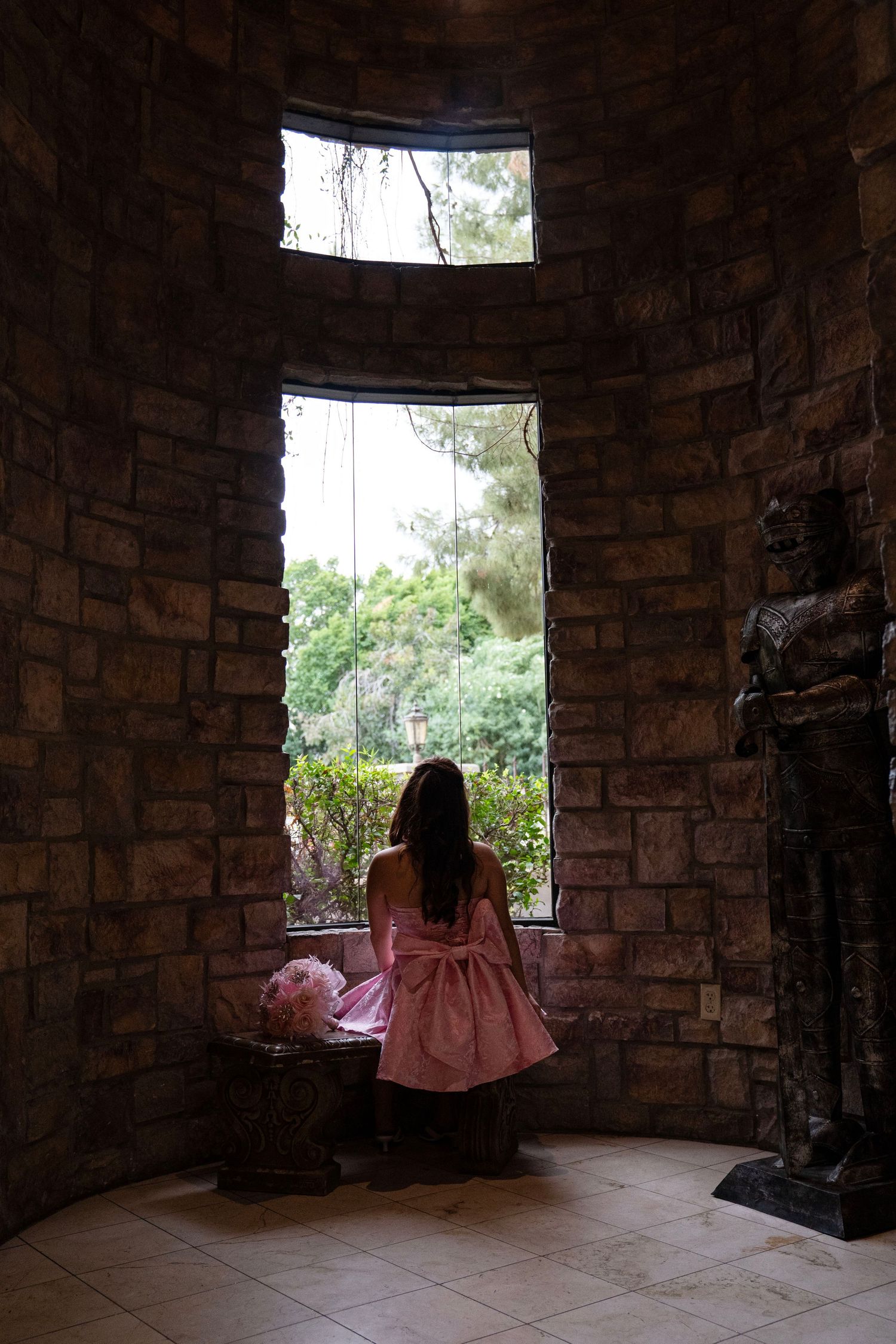 Silhouette of woman in pink bow dress standing at tall window in circular stone room.