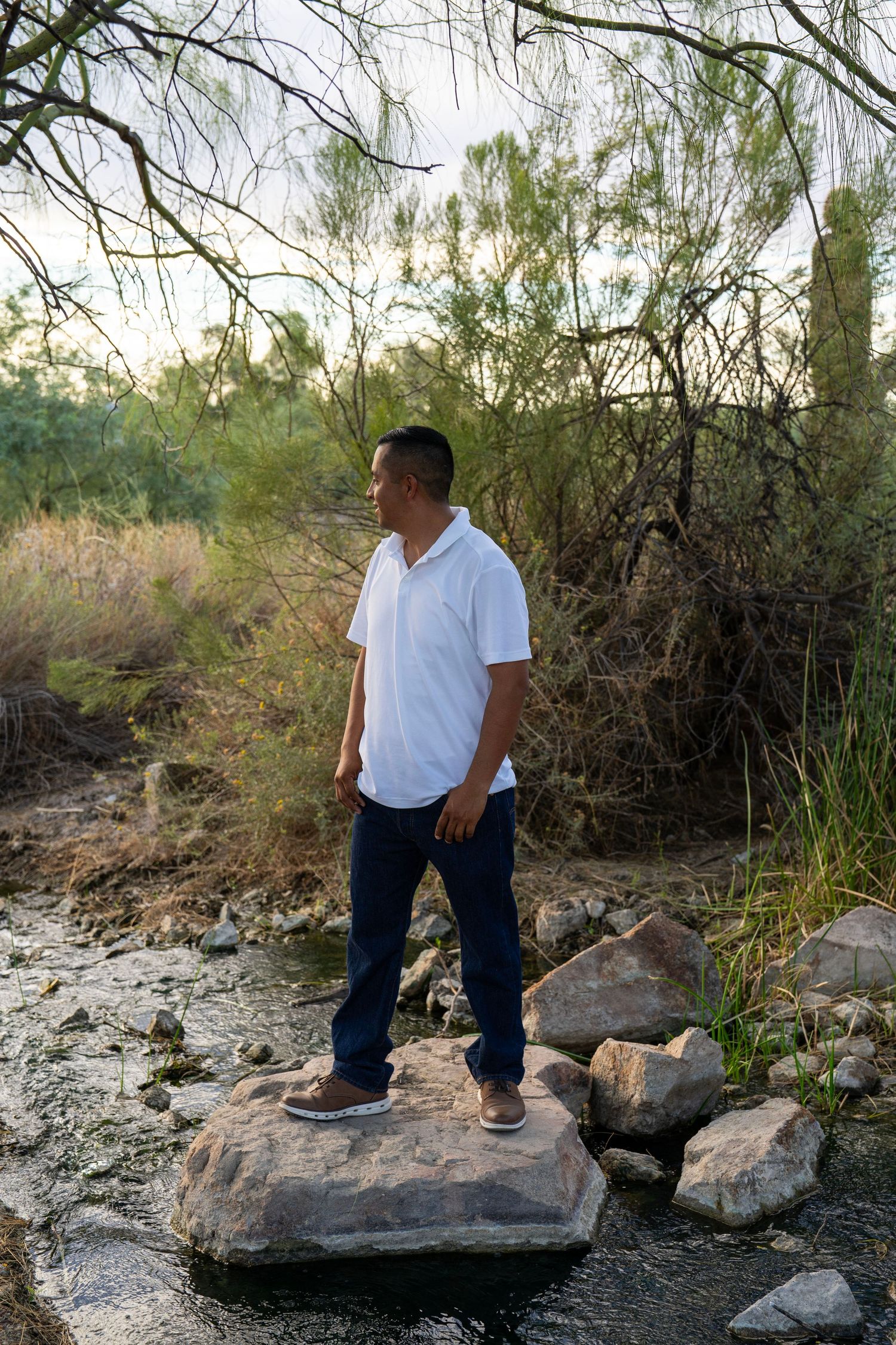 Man in white polo shirt and jeans stands on a rock by a stream, gazing into the distance amid lush greenery.