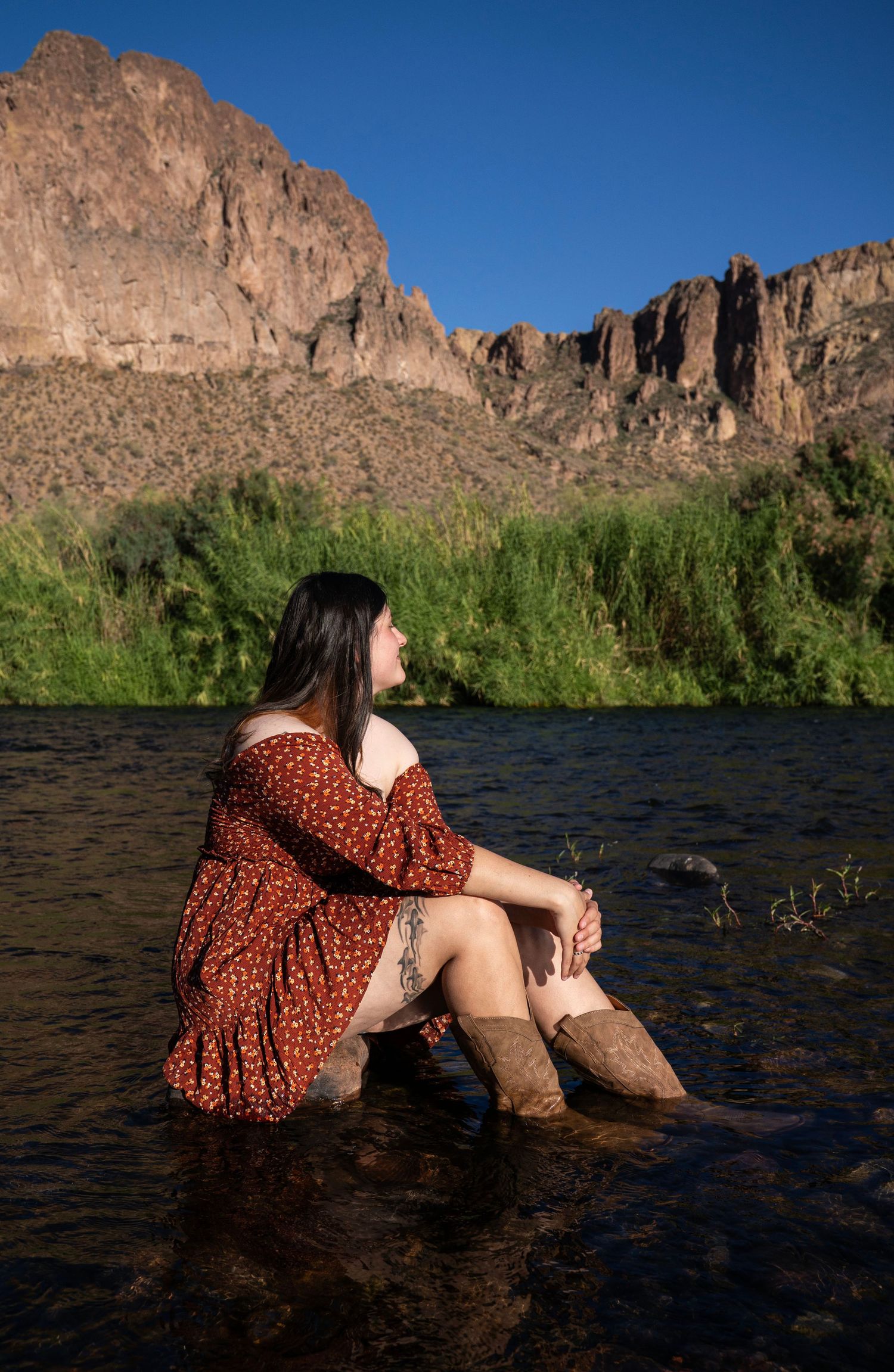 Woman in red floral dress and boots sitting in a river with rocky desert mountains in the background.