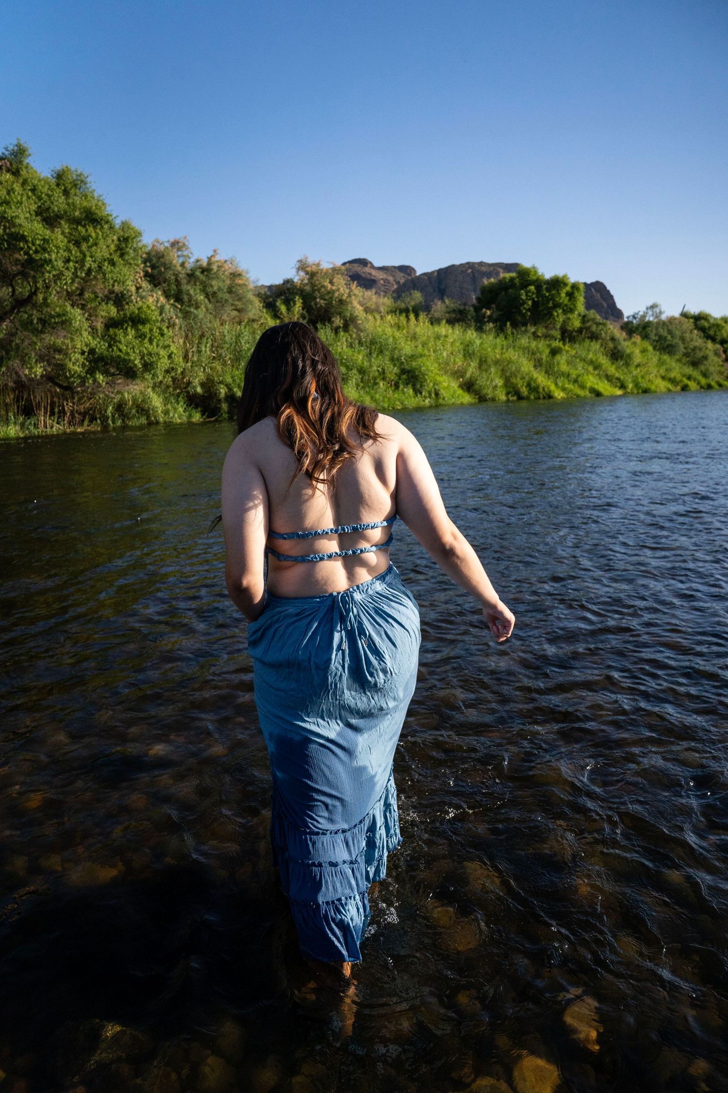 Woman in blue skirt wading into a calm river surrounded by lush green trees and rocky cliffs under a clear blue sky.