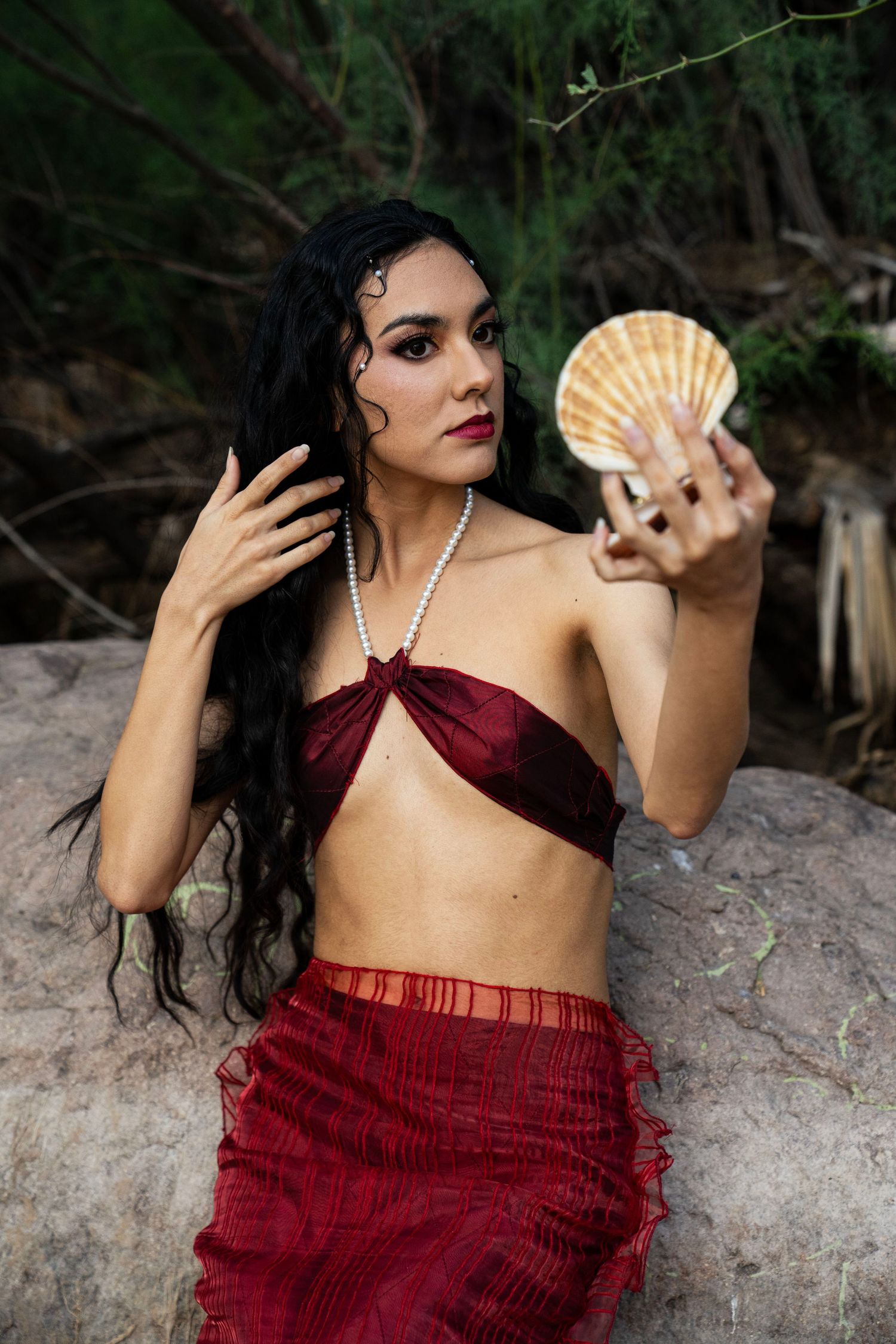 Woman in red mermaid costume holding a scallop shell while posing on rocks in a forest.