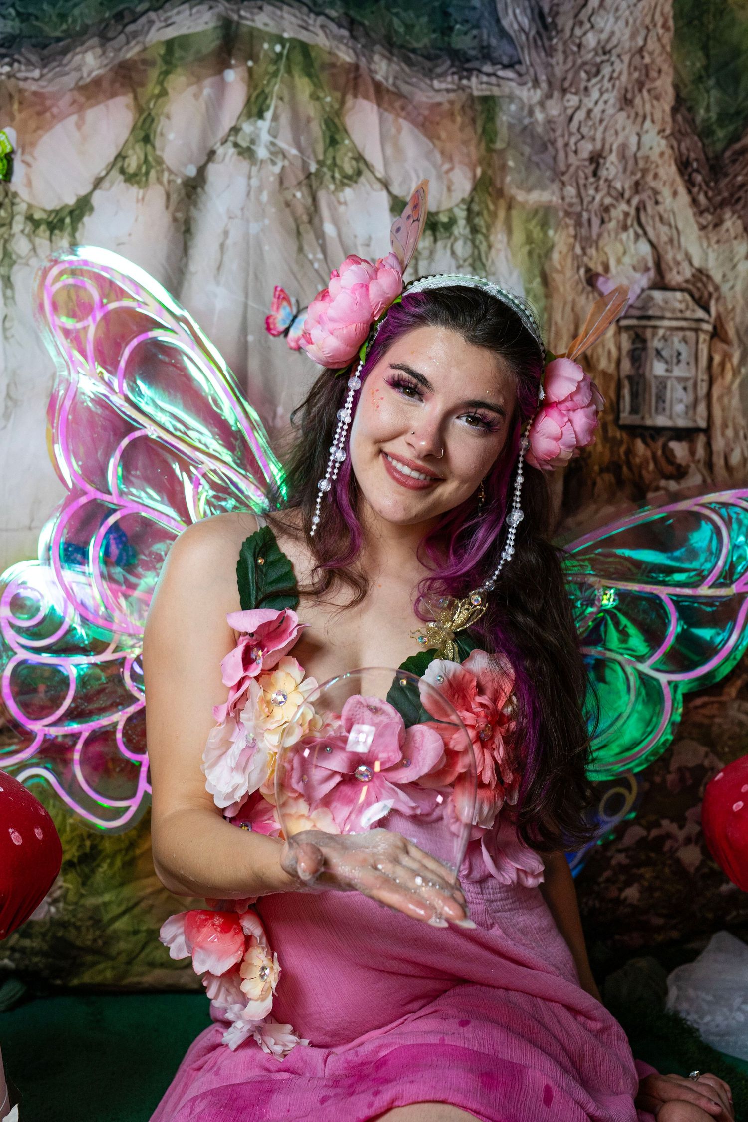 Fairy costume model with iridescent LED wings and pink floral dress poses in enchanted forest photo studio. Fantasy Photoshoot