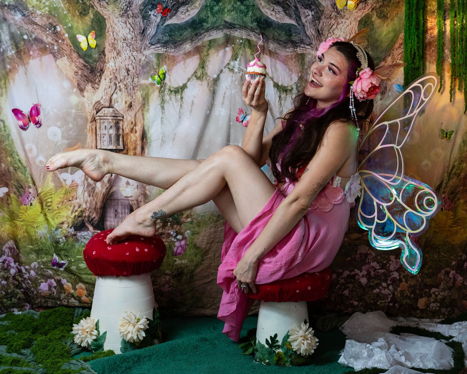 Woman in pink fairy costume with wings sits on red mushroom prop in enchanted forest setting holding cupcake. Fantasy Photoshoot