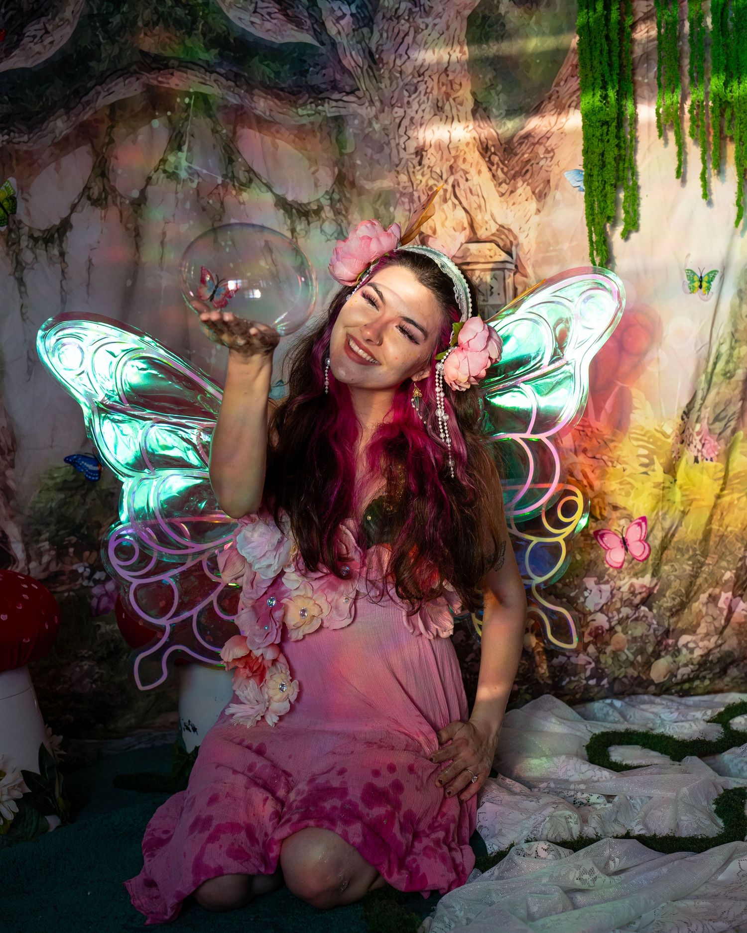 Joyful woman in pink fairy costume with LED wings holds a glass orb bubble in fantasy forest studio. Fantasy Photoshoot
