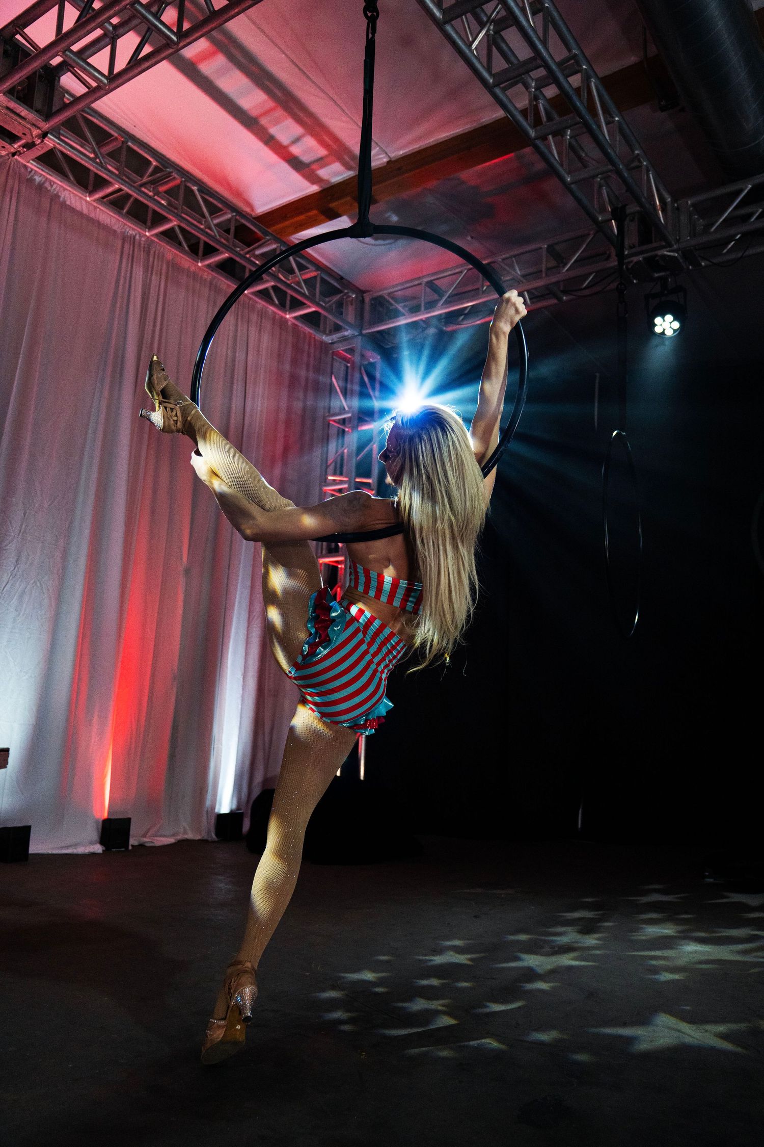 Rear view of blonde aerialist performing leg extension on aerial hoop under spotlights. Circus Photoshoot