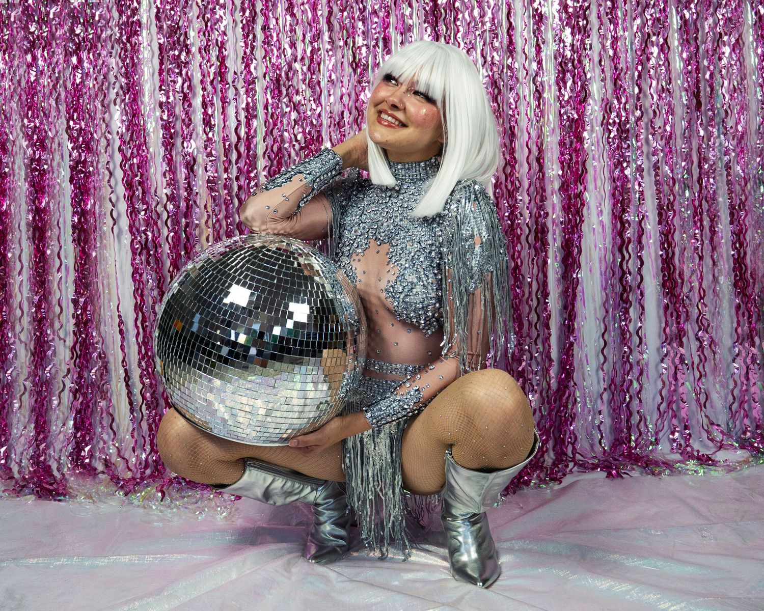 Smiling woman in silver sequin fringe outfit holds large disco ball, wearing white wig, pink backdrop.