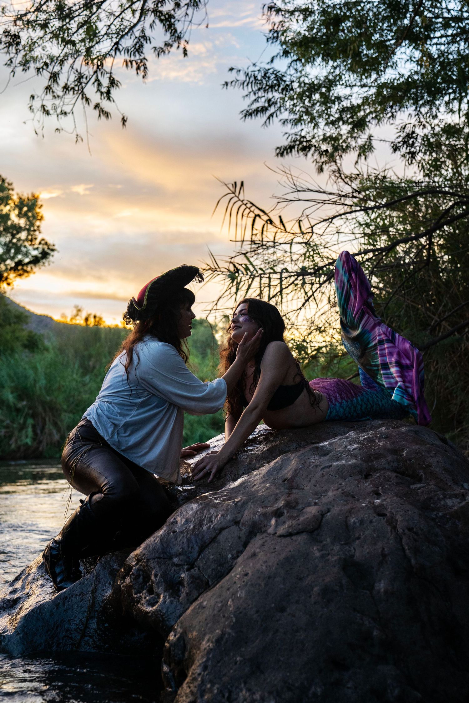 Two women in pirate and mermaid costumes sitting together on rocks by river at dusk. Mermaid photoshoot, fantasy photoshoot.