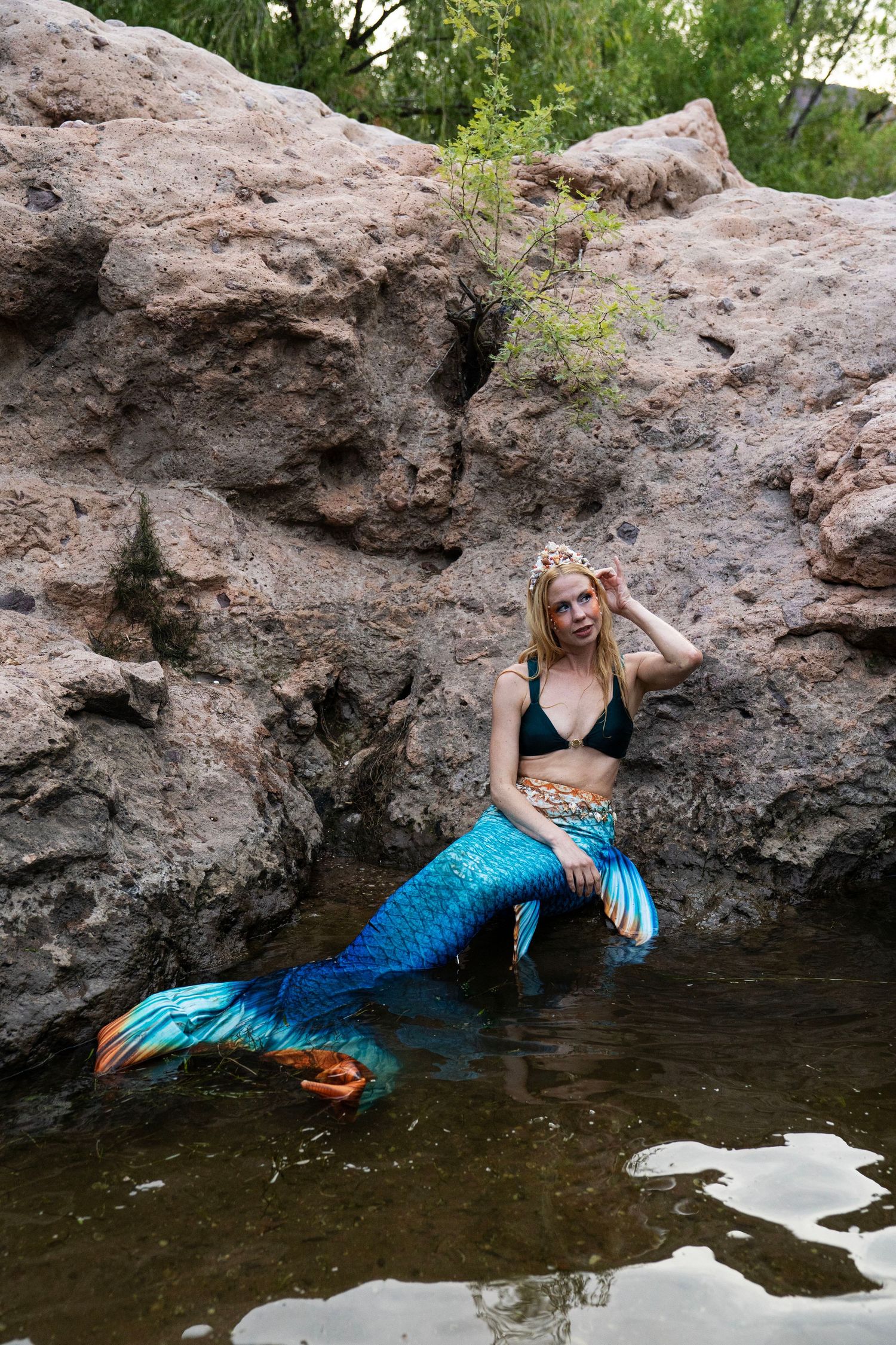 A blonde woman wearing a blue mermaid tail and black bikini top sits against rocky cliffs. Fantasy Photoshoot