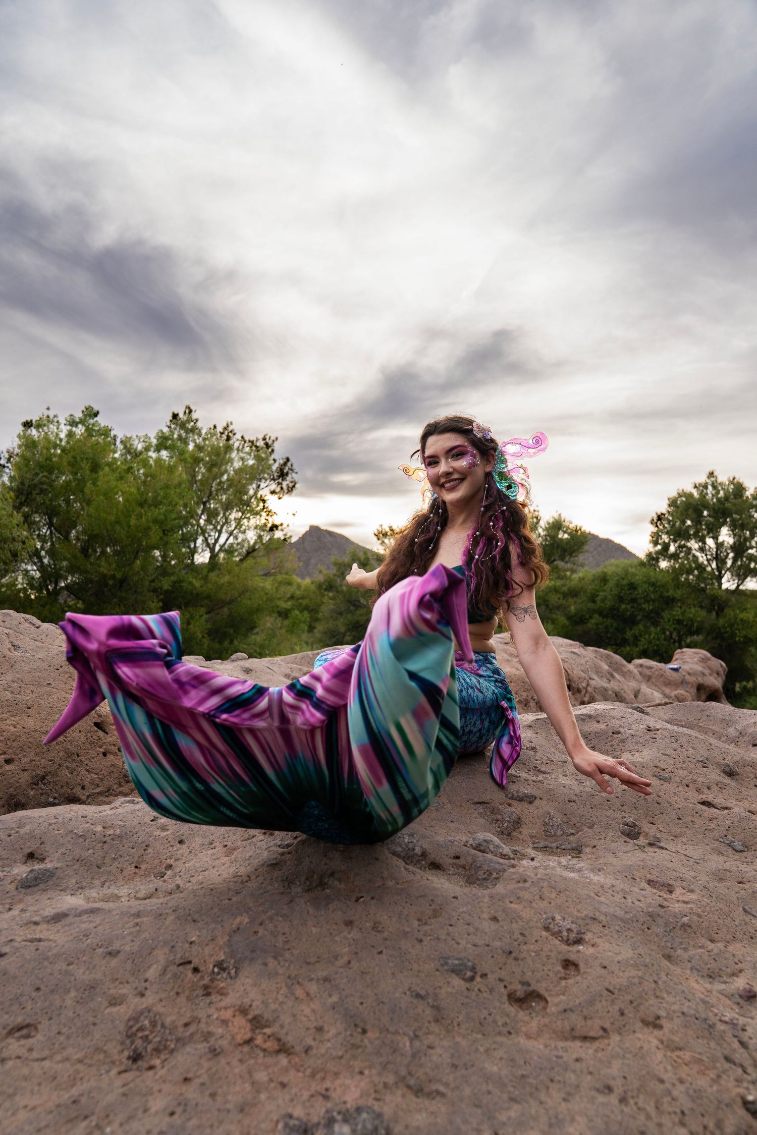 Woman in a flowing pink and teal mermaid tail costume posing dramatically on rocky terrain under cloudy skies. Fantasy Photoshoot