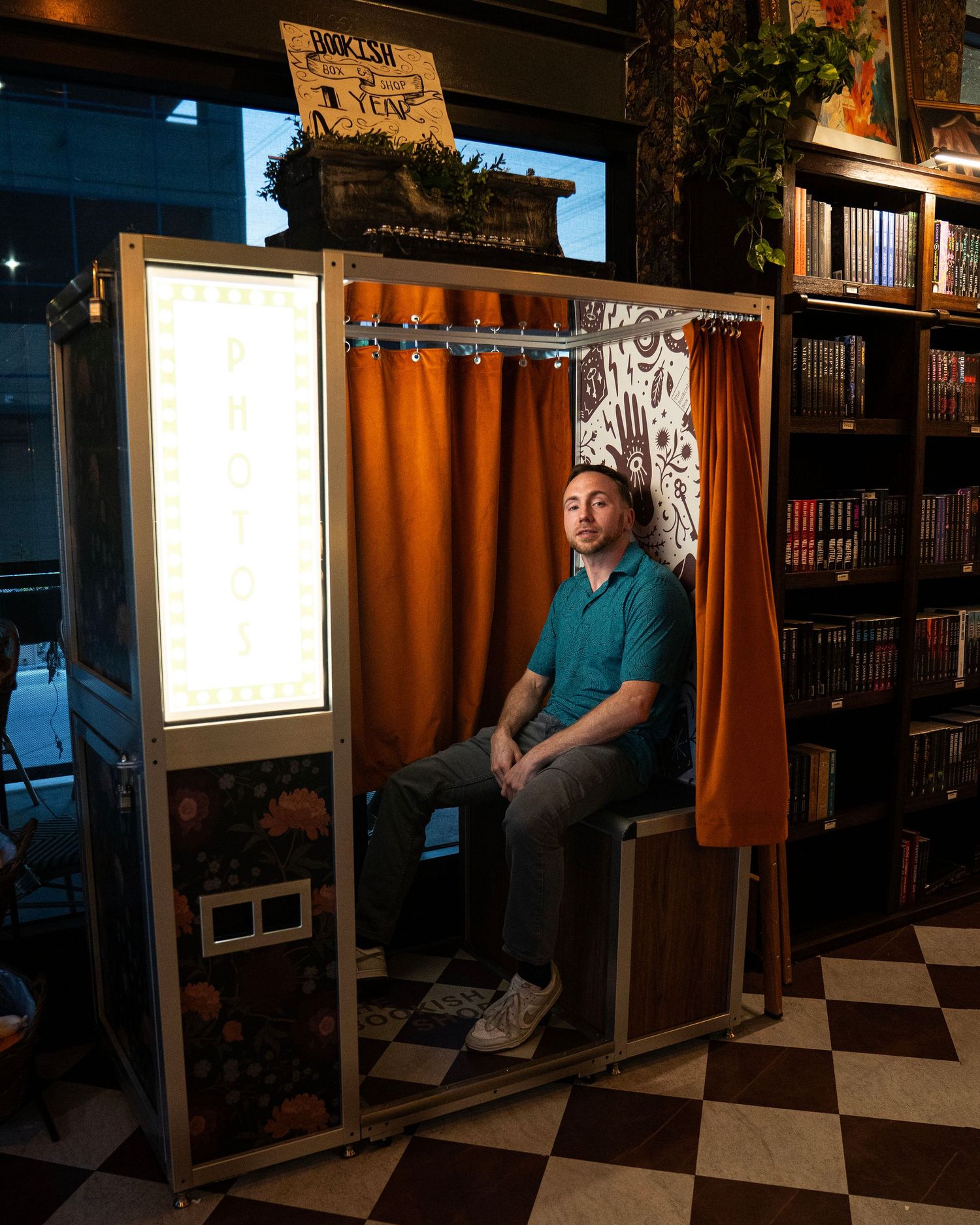 Man sitting inside a vintage photo booth with orange curtains in a cozy bookstore setting.