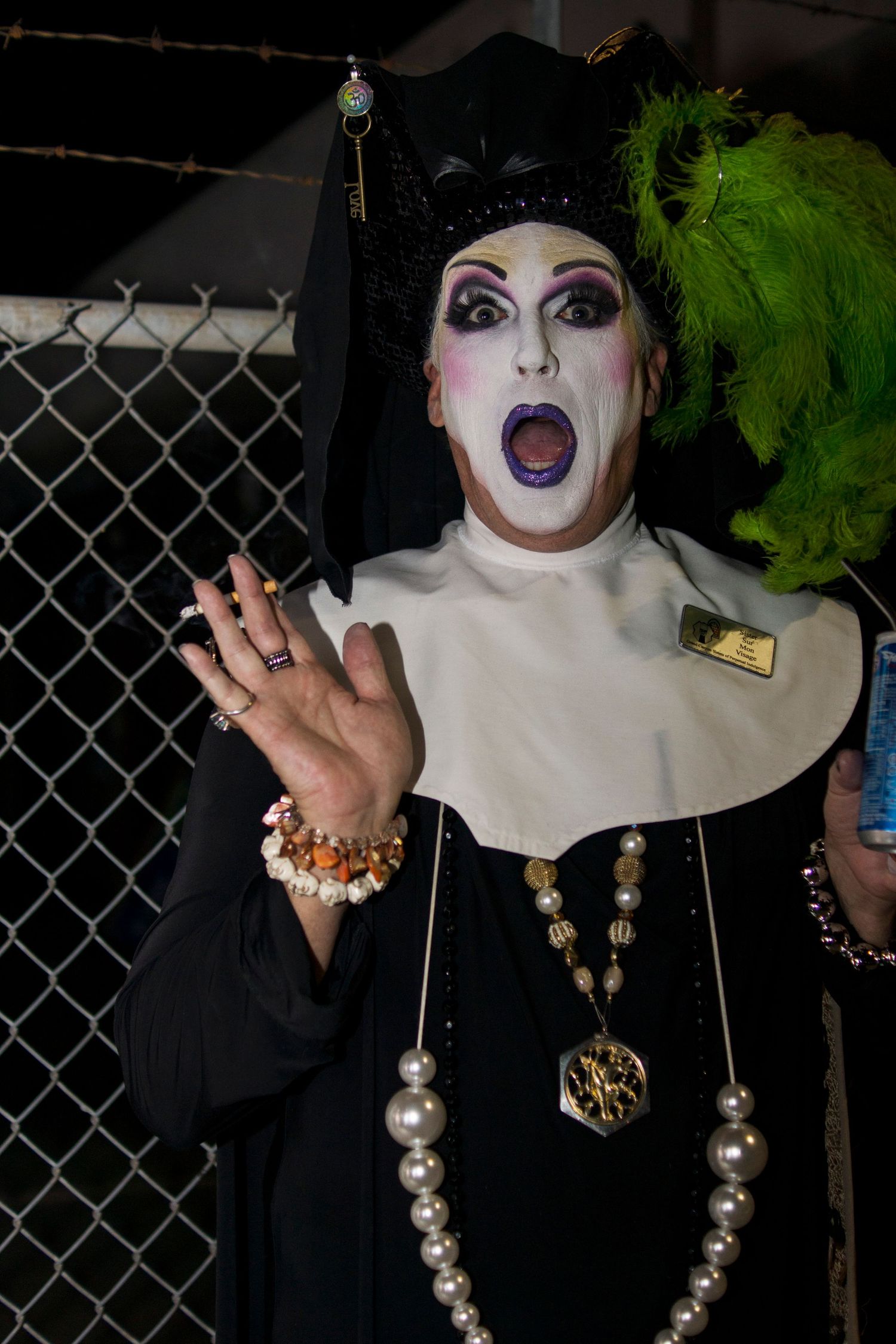 Drag performer dressed as a nun with white face paint, green feather wig, pearl necklaces, and dramatic makeup, waving at camera.
