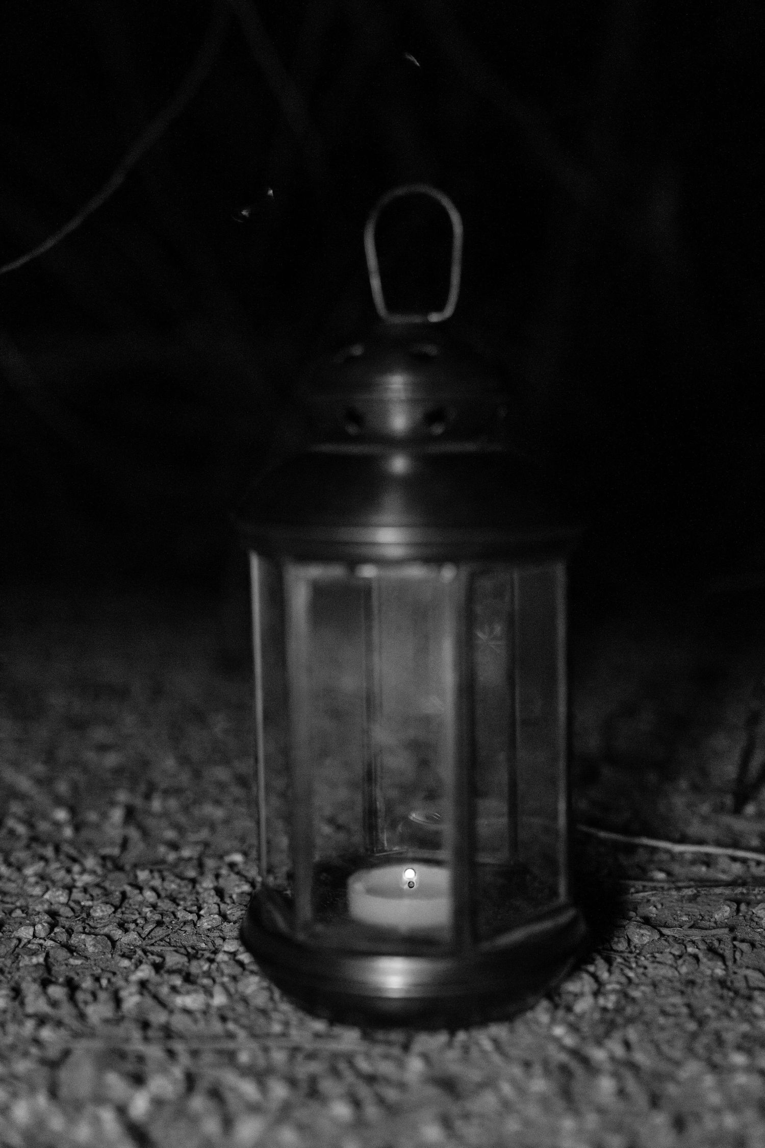 Black and white photo of a small lantern with a lit candle glowing inside, resting on gravel in the dark.