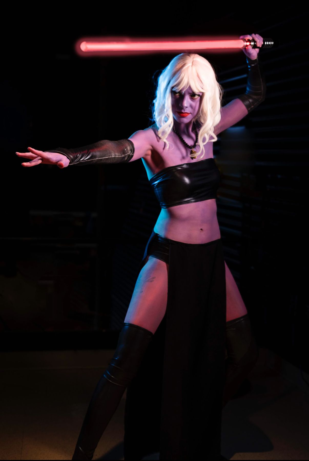 Woman cosplaying as a Sith warrior in black costume holding a glowing red lightsaber in a dramatic pose.