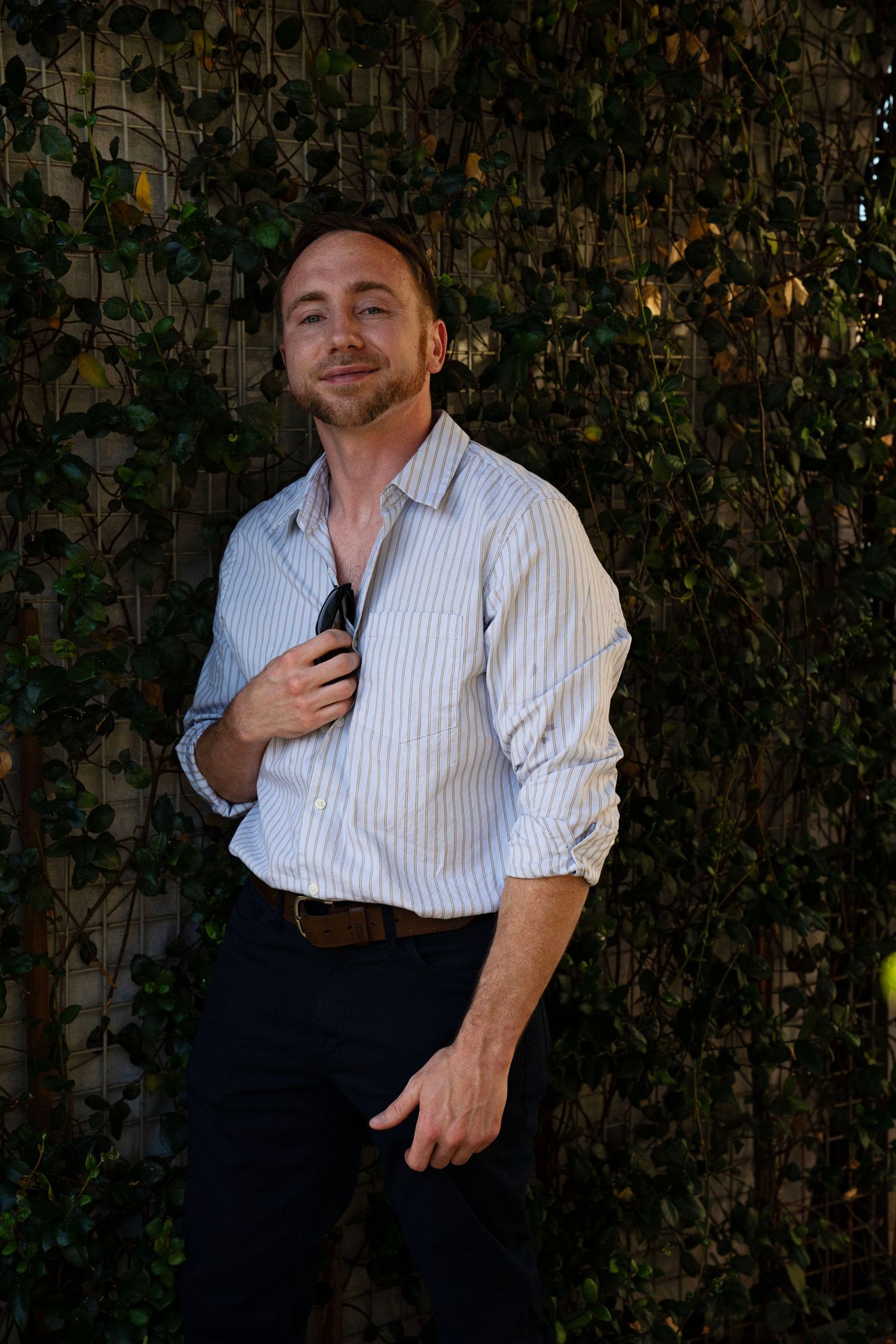 Man in striped shirt holding sunglasses poses against ivy-covered wall outdoors.