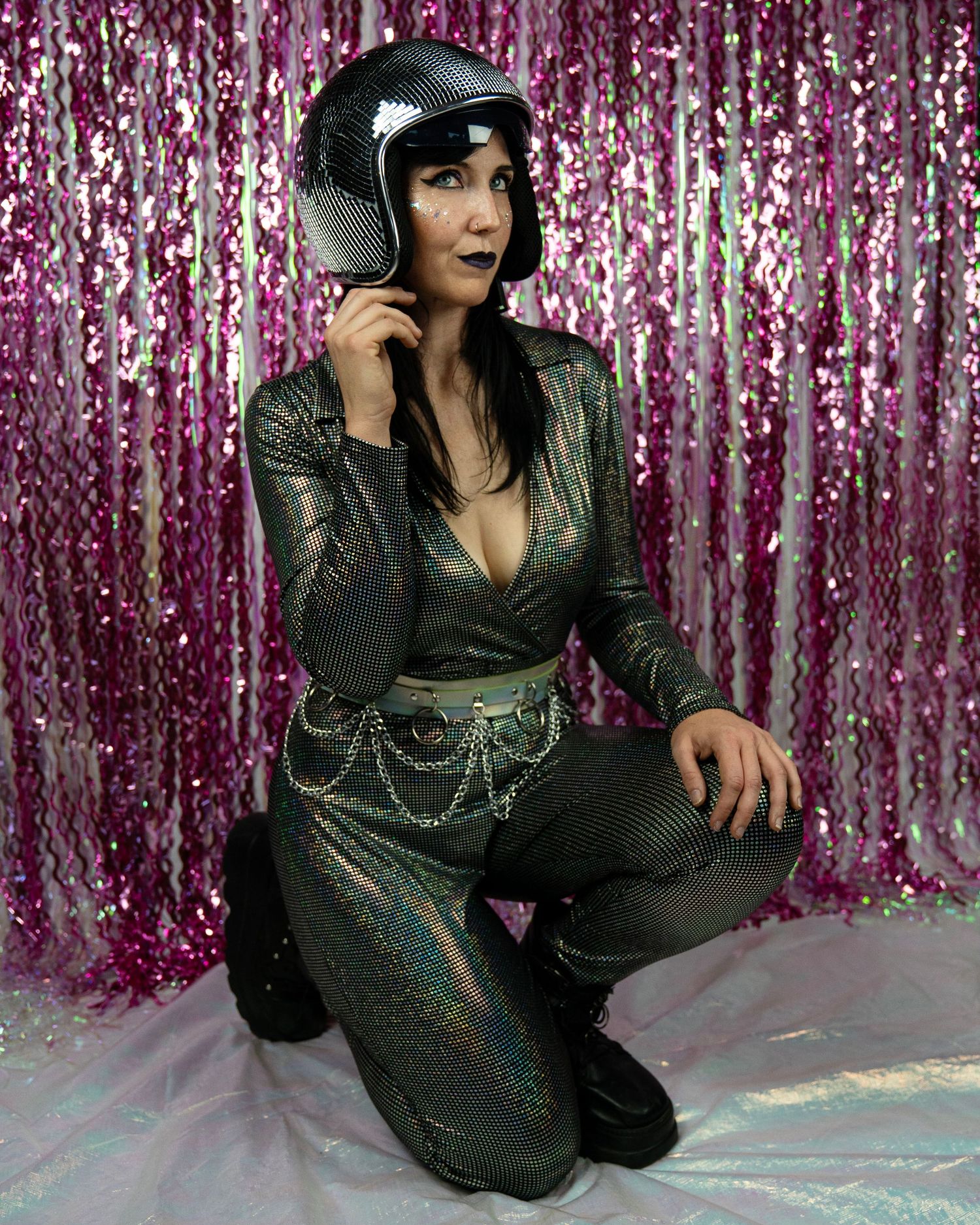 A woman in a holographic dark jumpsuit kneels while wearing a disco ball helmet against pink tinsel.