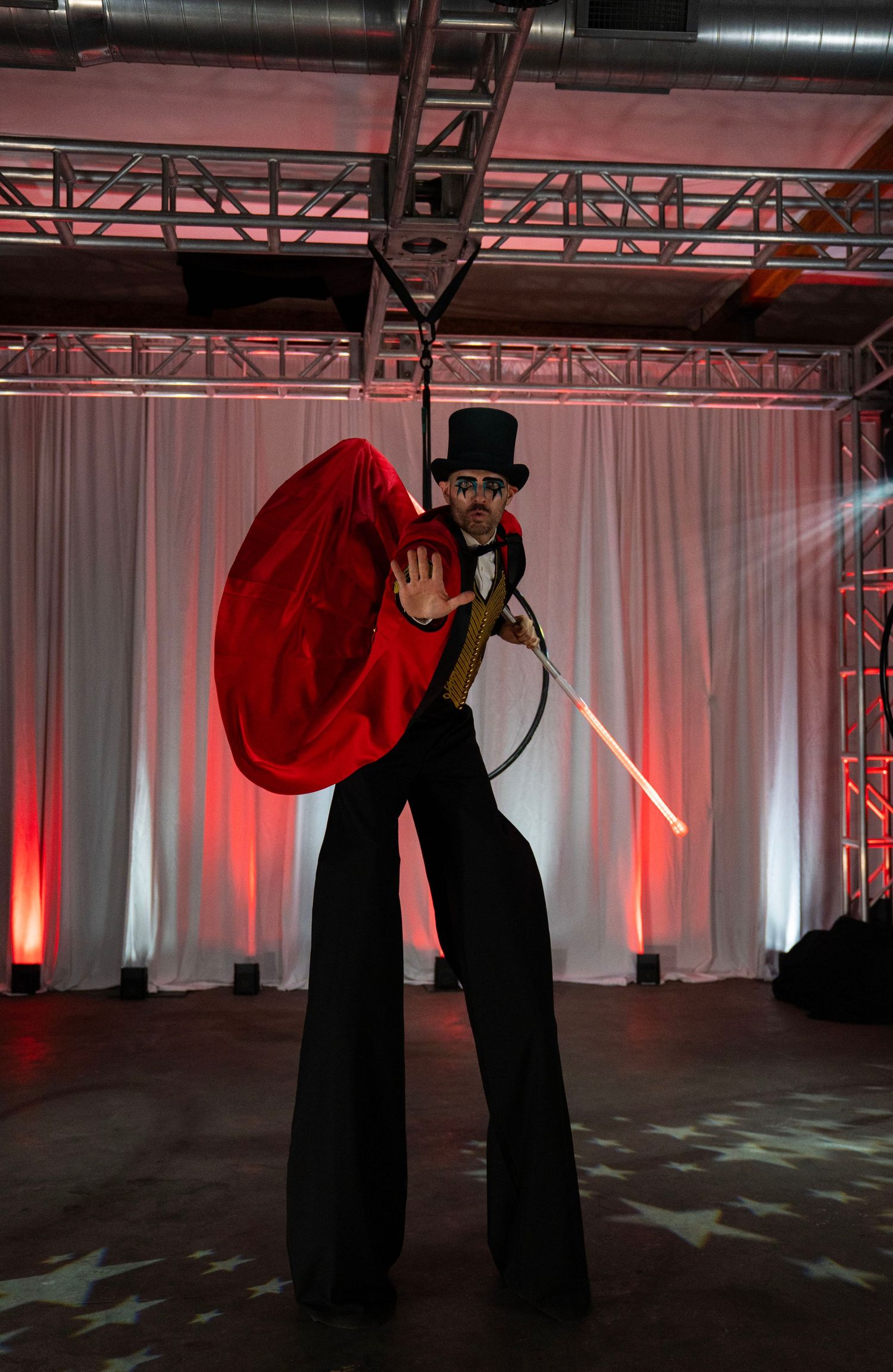 Circus ringmaster stilt walker in top hat holds red cape and glowing sword at dramatic event.
