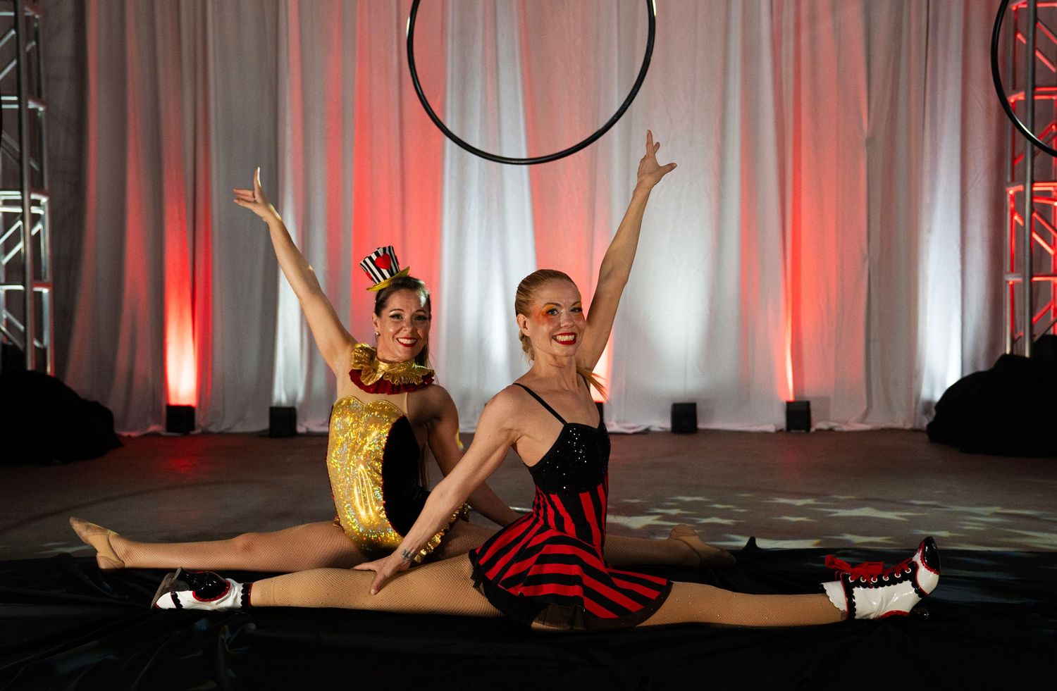 Two smiling acrobats in circus costumes posing in splits with aerial hoop backdrop.