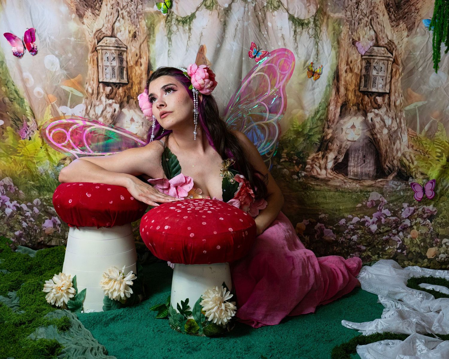 Fairy costume model with iridescent wings and pink flowers poses with red toadstools in enchanted woodland backdrop. Fantasy Photoshoot