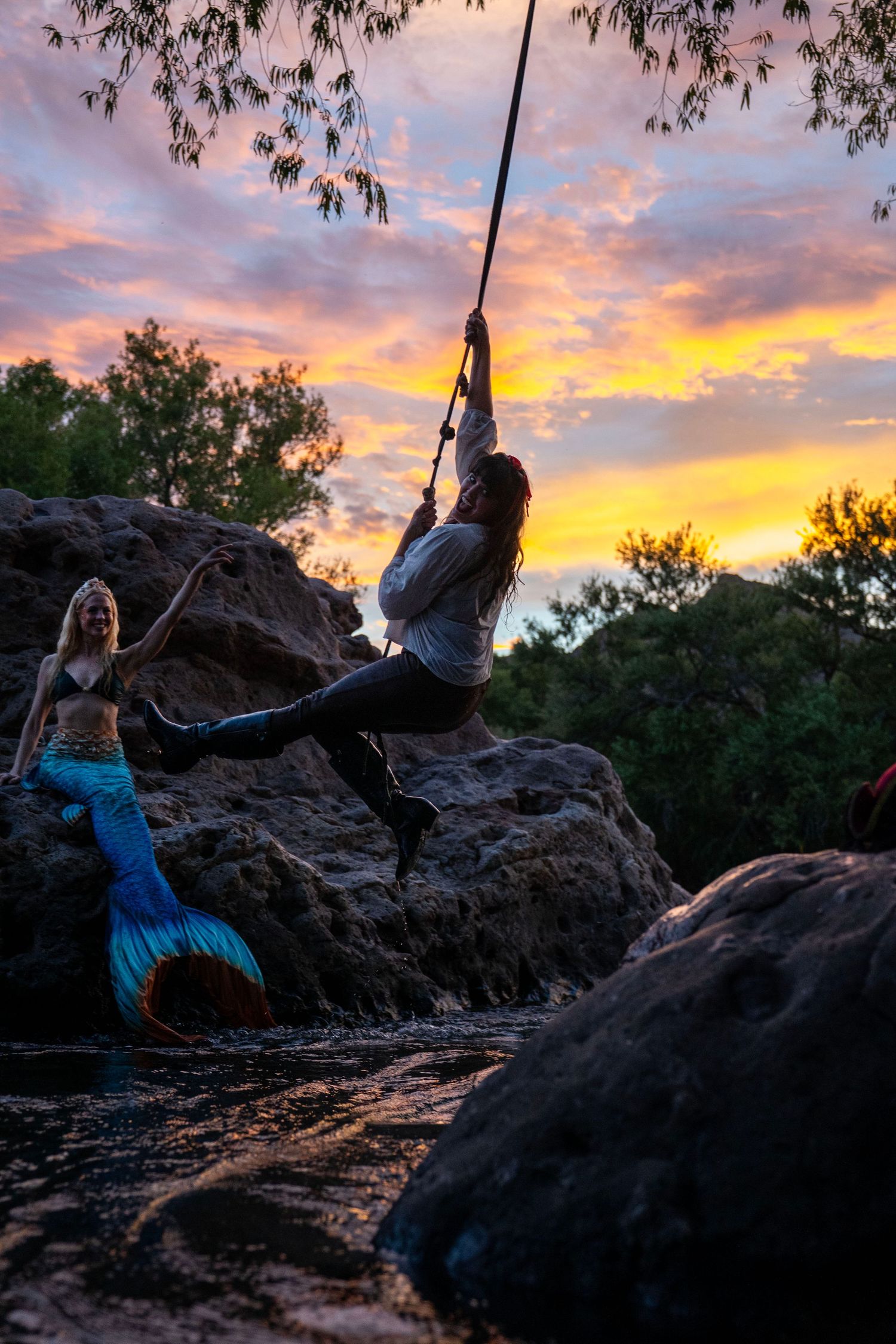 A person swings on a rope while a mermaid watches from rocks against a vivid colorful sunset sky. Mermaid photoshoot, fantasy photoshoot.